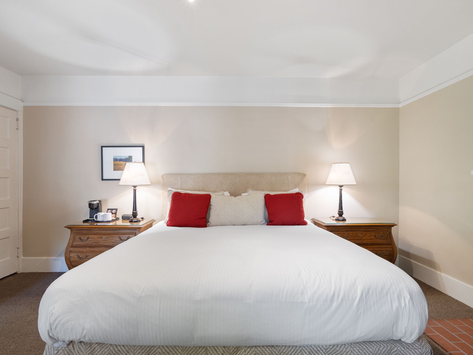 A hotel bed with a white comforter, two red and two white pillows, bedside tables with lamps, and a framed picture.