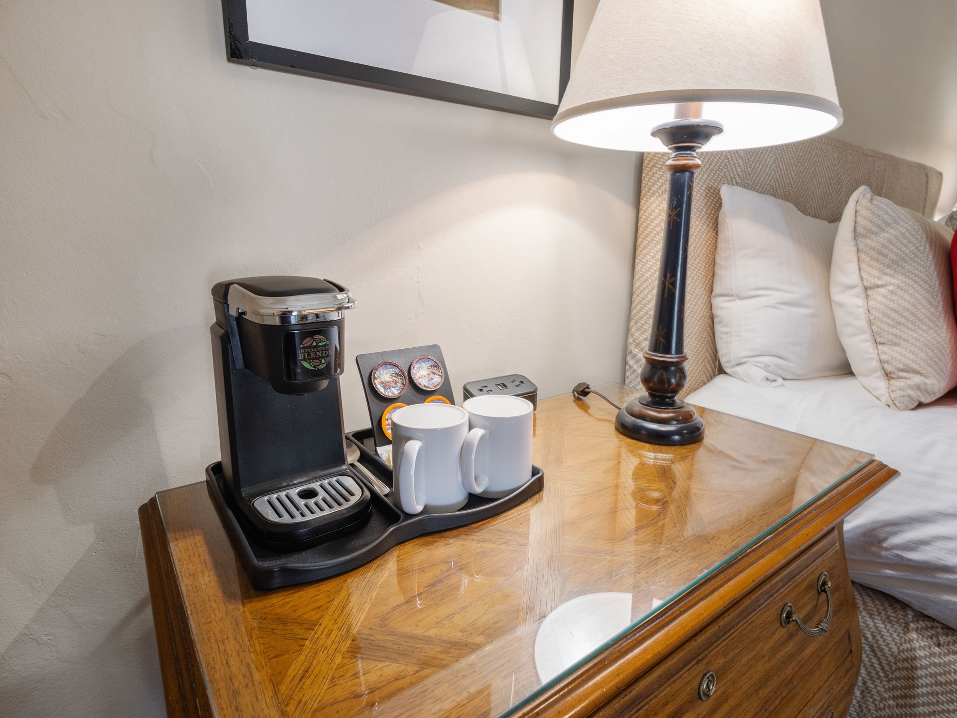 A Keurig coffee maker and two white mugs sit on a wooden hotel nightstand next to a lamp and a bed.