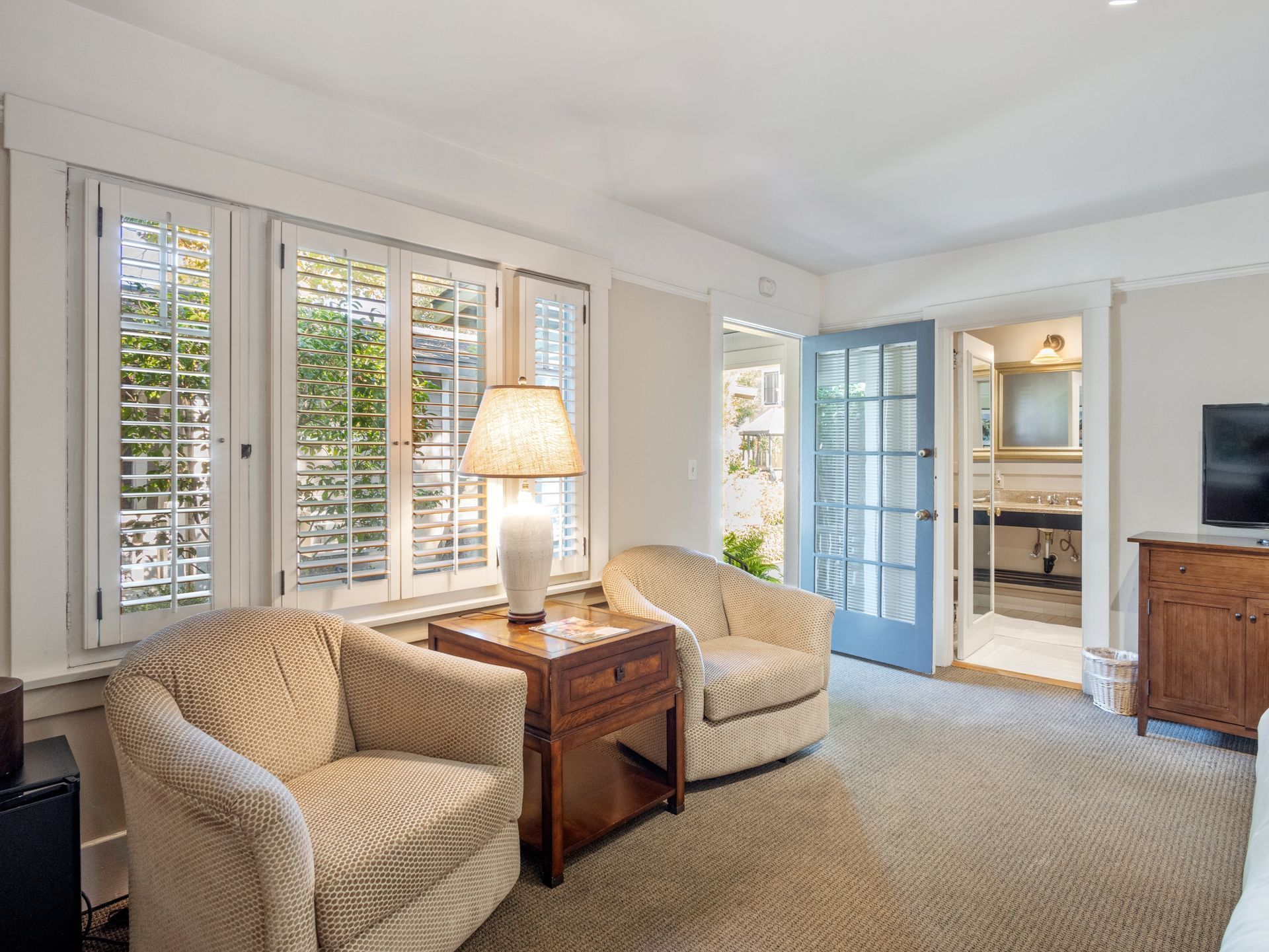 A cozy room with two beige armchairs, a wooden side table, a lamp, white shutters, and a blue door leading to a bathroom.