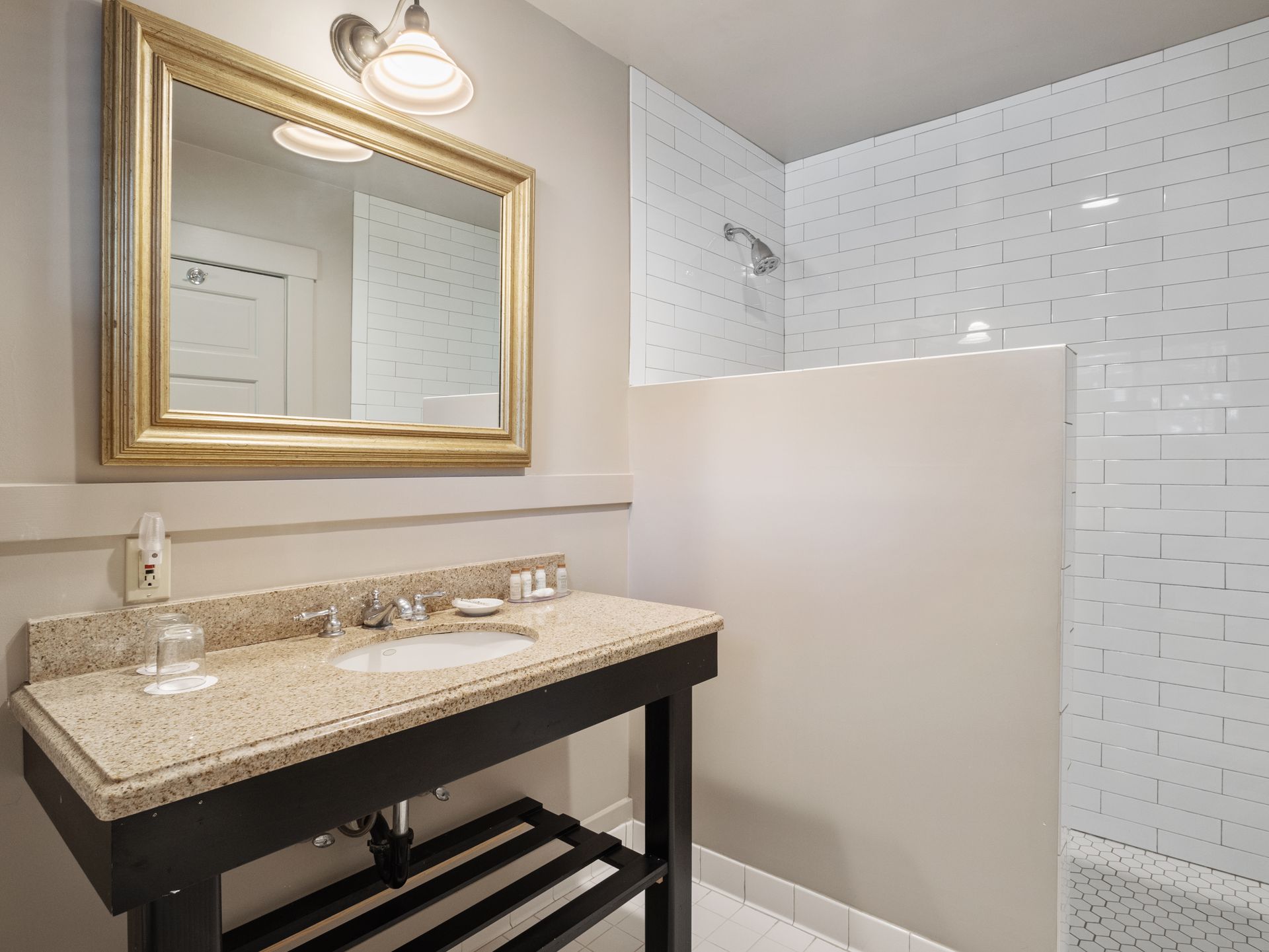 A bathroom vanity with a beige granite countertop, dark base, gold-framed mirror, and a tiled shower stall to the right.