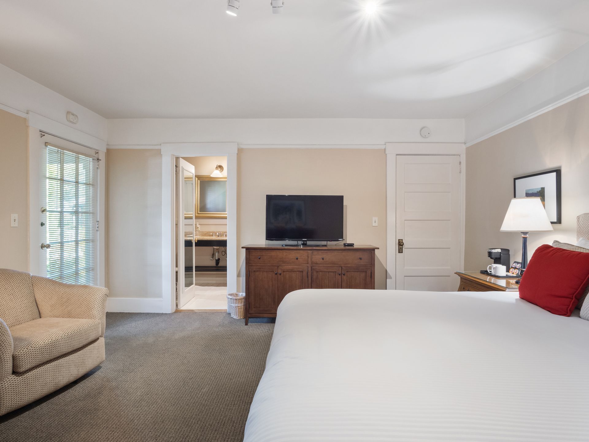 A hotel room featuring a white bed with red pillows, a wooden dresser with a TV, a corner armchair, and an open bathroom.