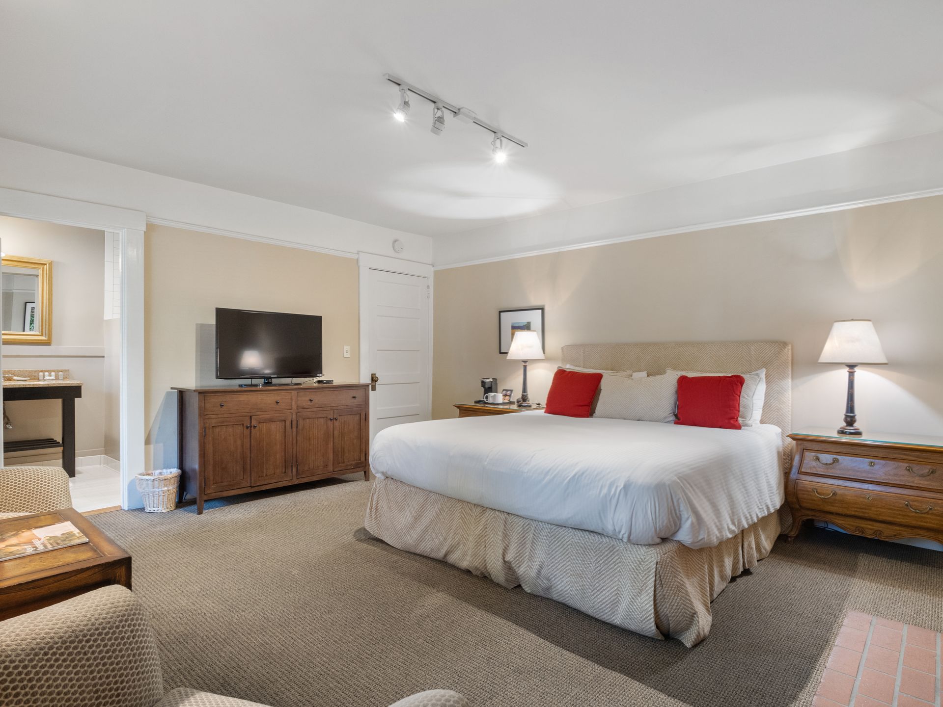 A hotel room with a king bed featuring red pillows, a wooden dresser with a TV, nightstands, and a corner bathroom view.
