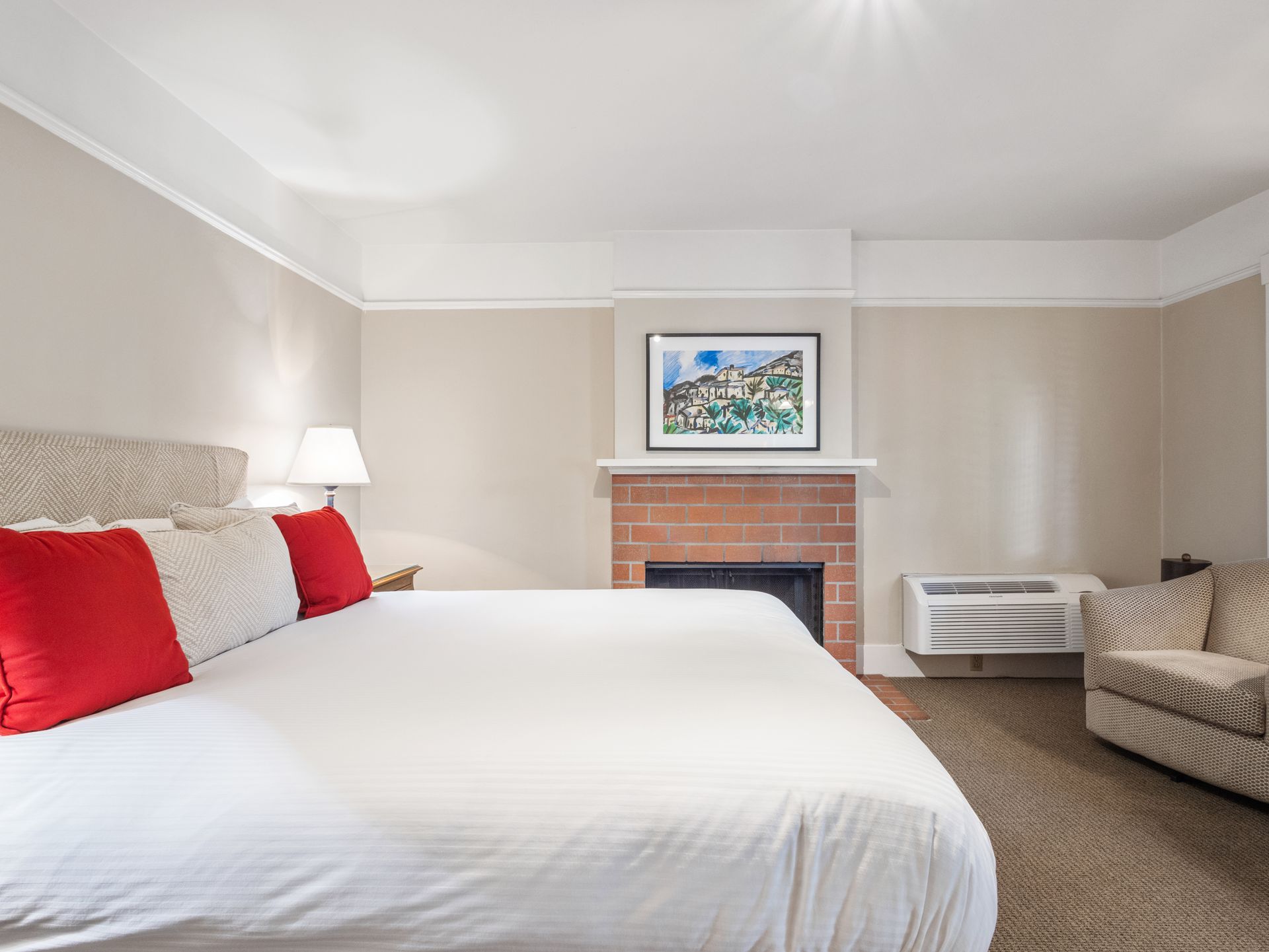 A hotel bedroom with a large bed, red pillows, a brick fireplace, a small armchair, and an air conditioning unit.