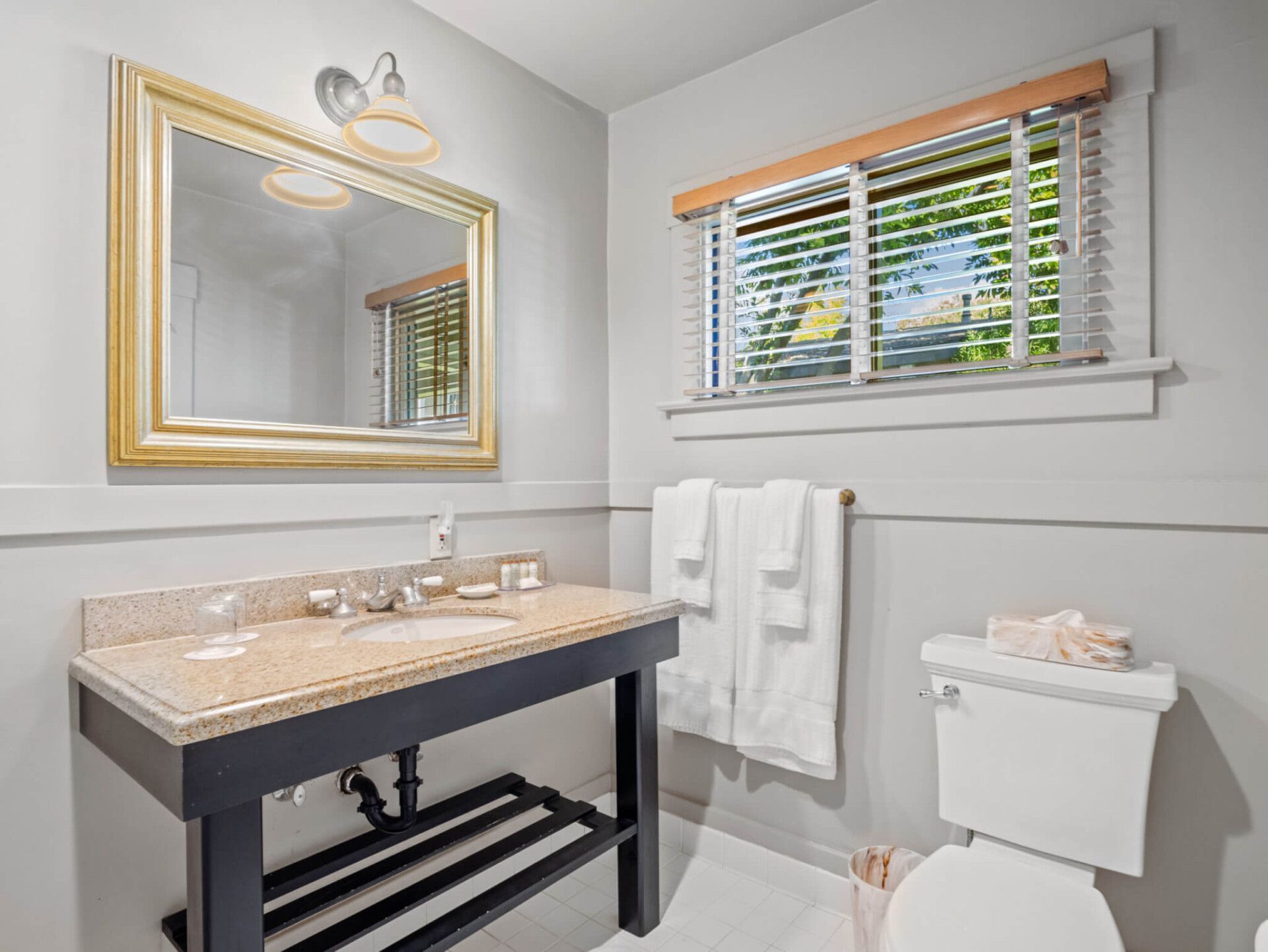 A bathroom with a black vanity, granite countertop, gold-framed mirror, and a toilet with towels hung beside it.