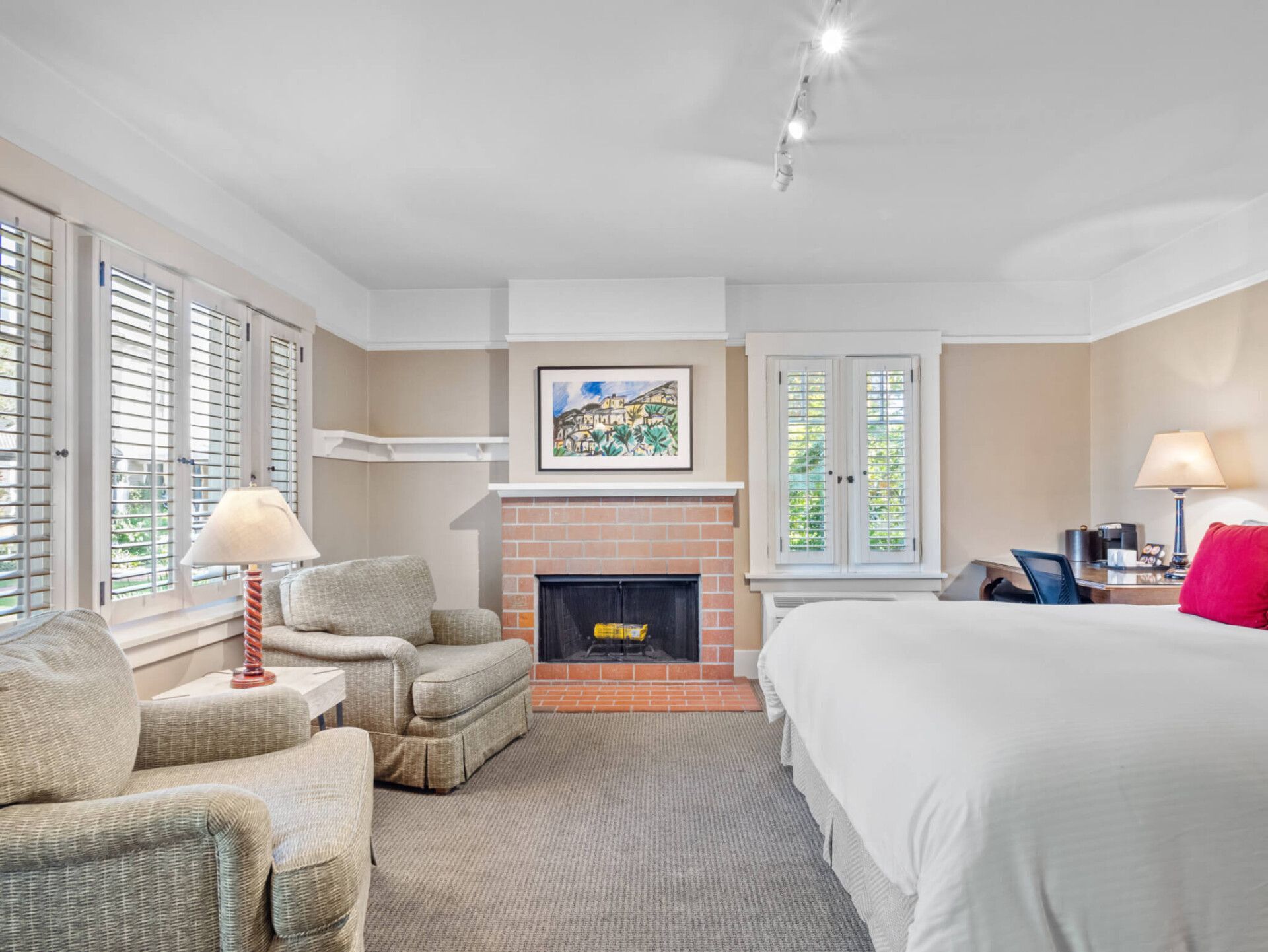 A hotel room features a large bed with white bedding, two cozy beige armchairs, a brick fireplace, and shuttered windows.