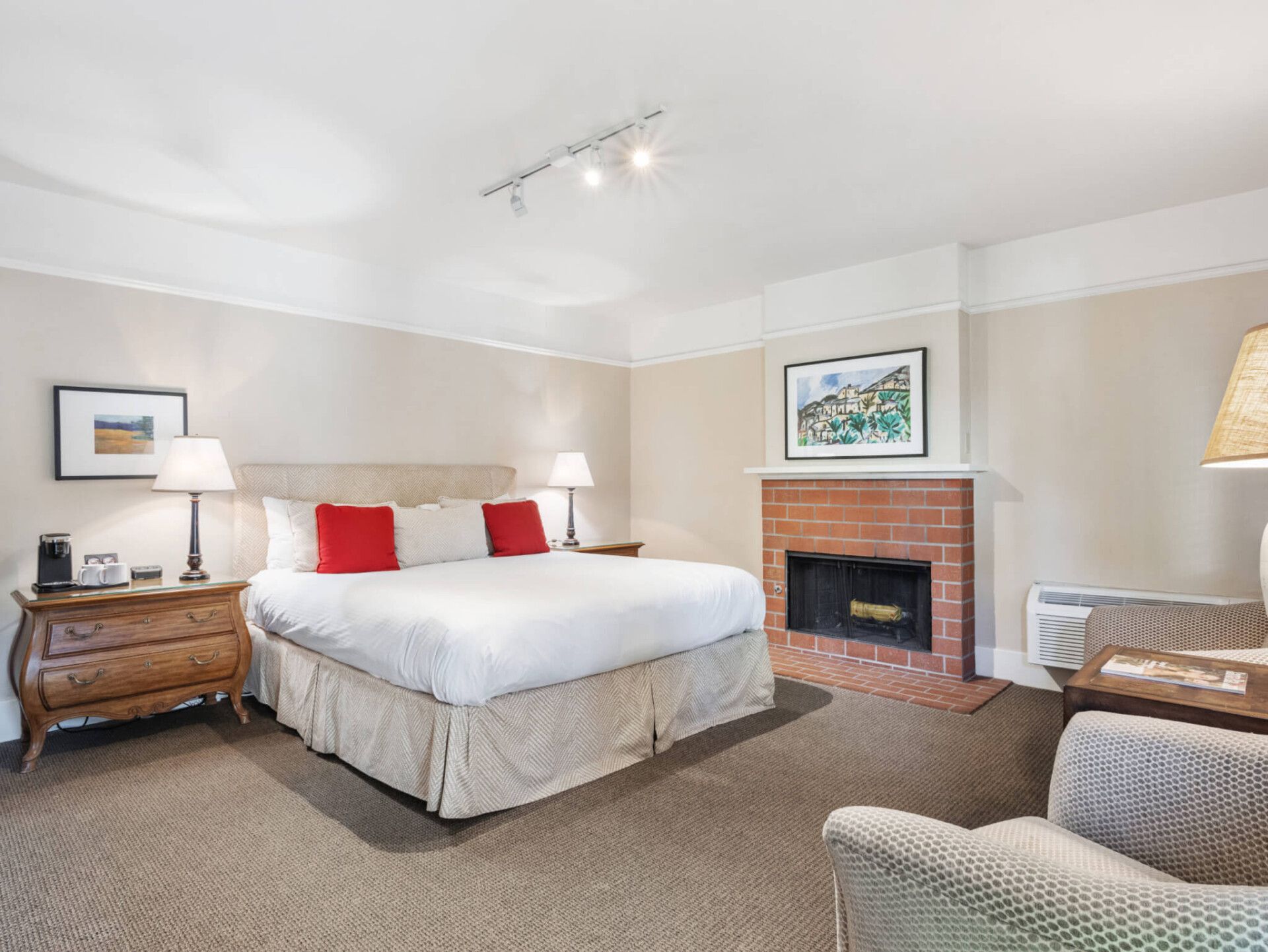 A cozy hotel room featuring a king bed with red pillows, a brick fireplace, a wooden dresser, and two armchairs.