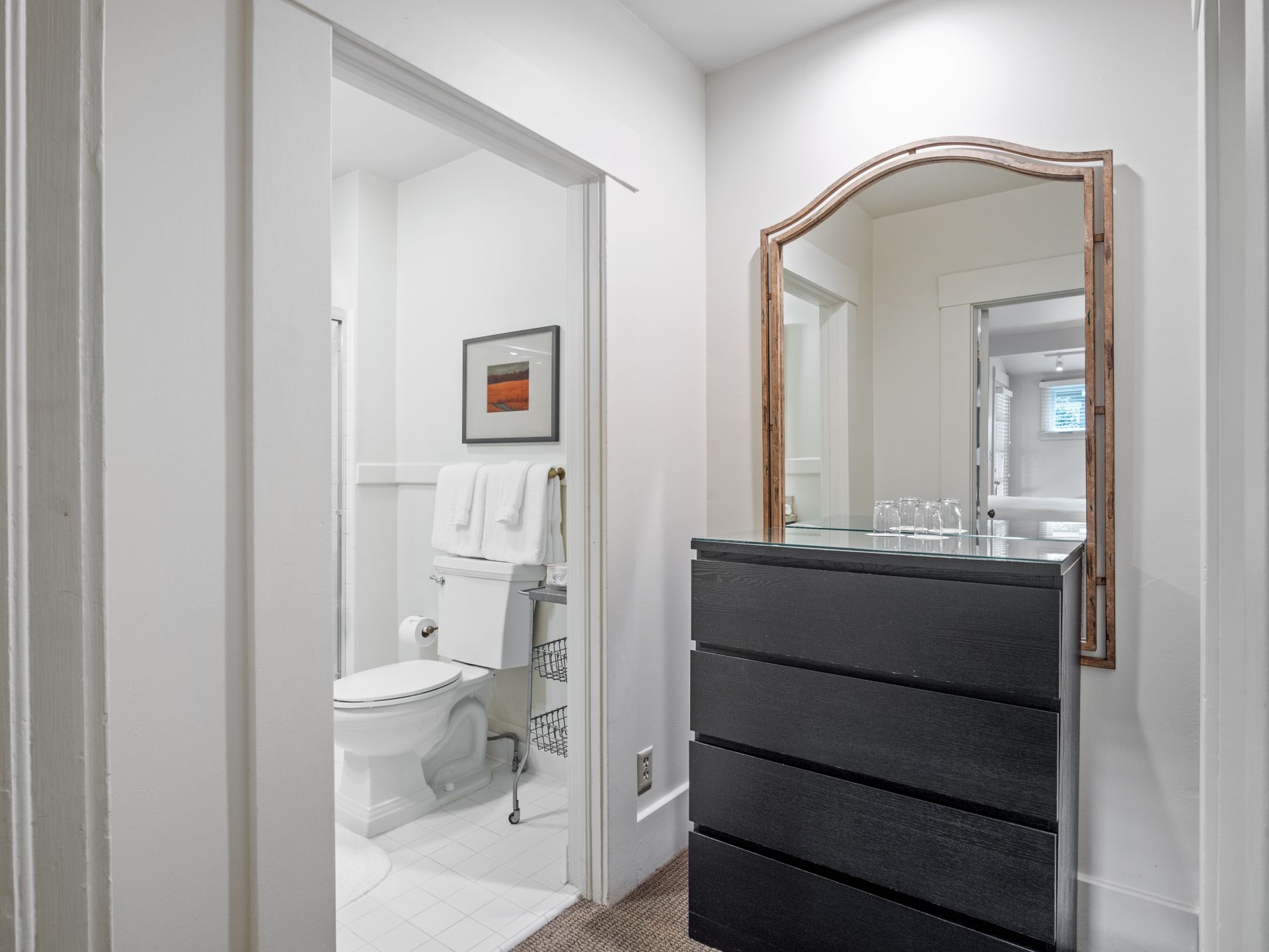 A view into a white bathroom with a toilet and hanging towels, next to a black four-drawer dresser with a wooden mirror.