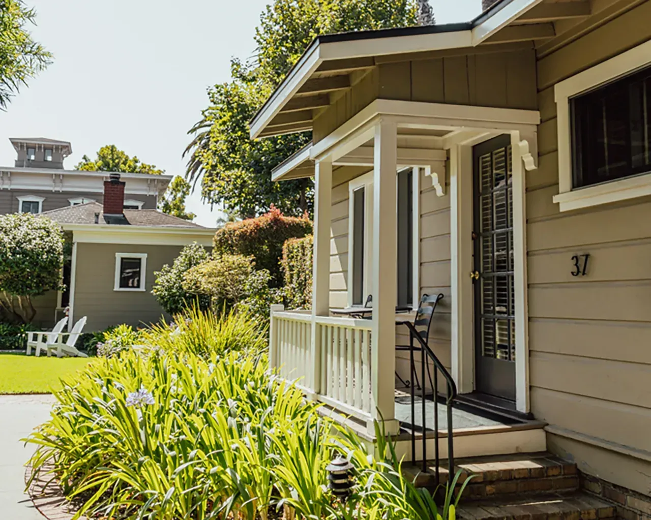 The exterior of a tan cottage with a small front porch, surrounded by lush green garden plants on a sunny day.