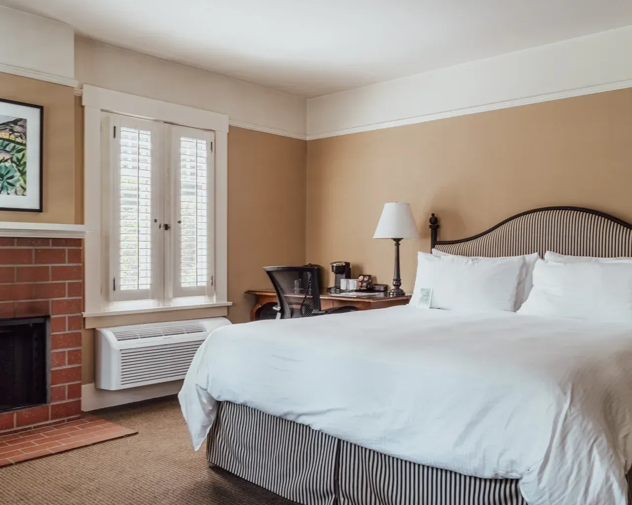 A hotel room with a large bed, striped headboard, fireplace, and a window with white shutters.