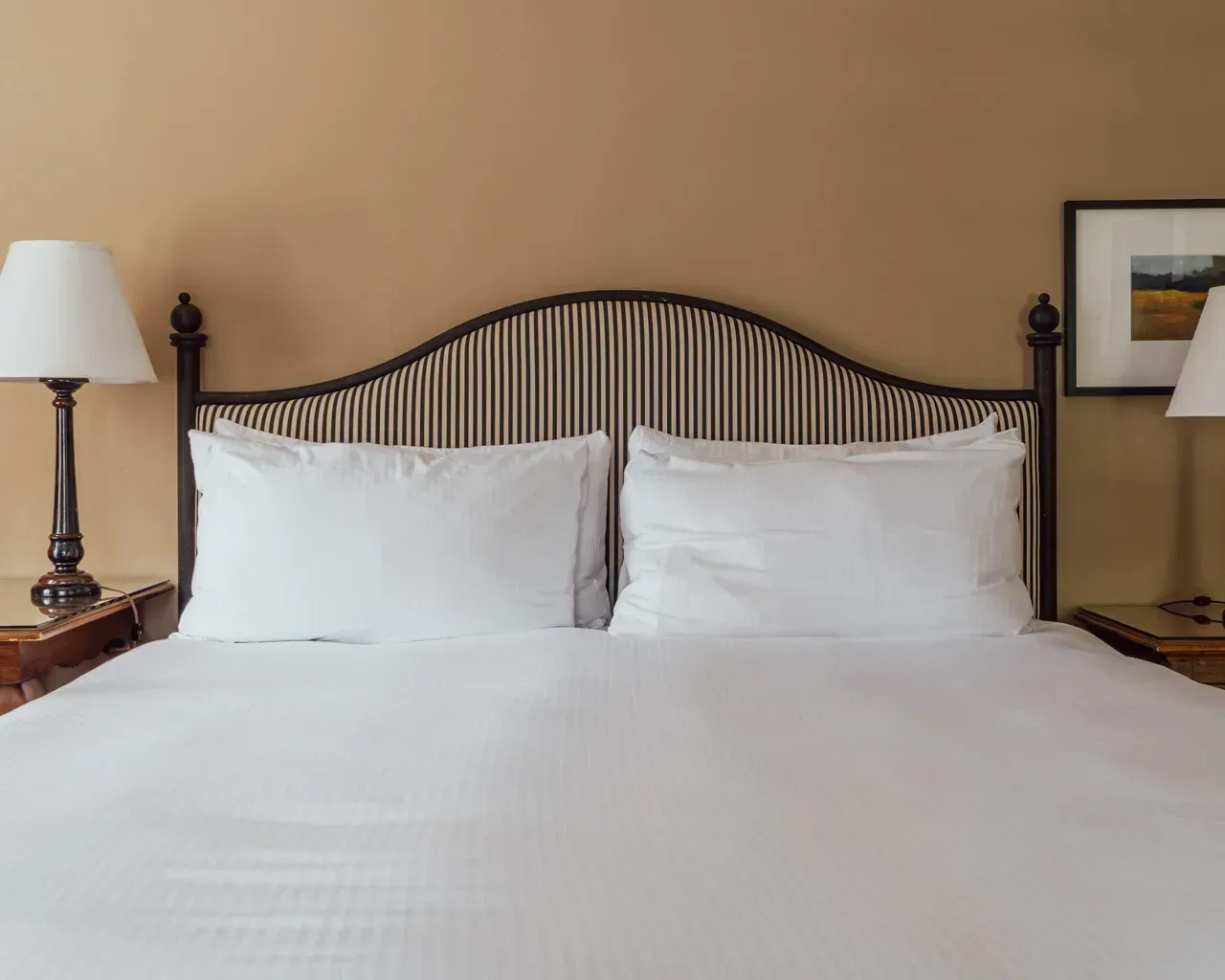 A neatly made bed with a striped headboard, white pillows, lamps on matching bedside tables, and framed wall art.