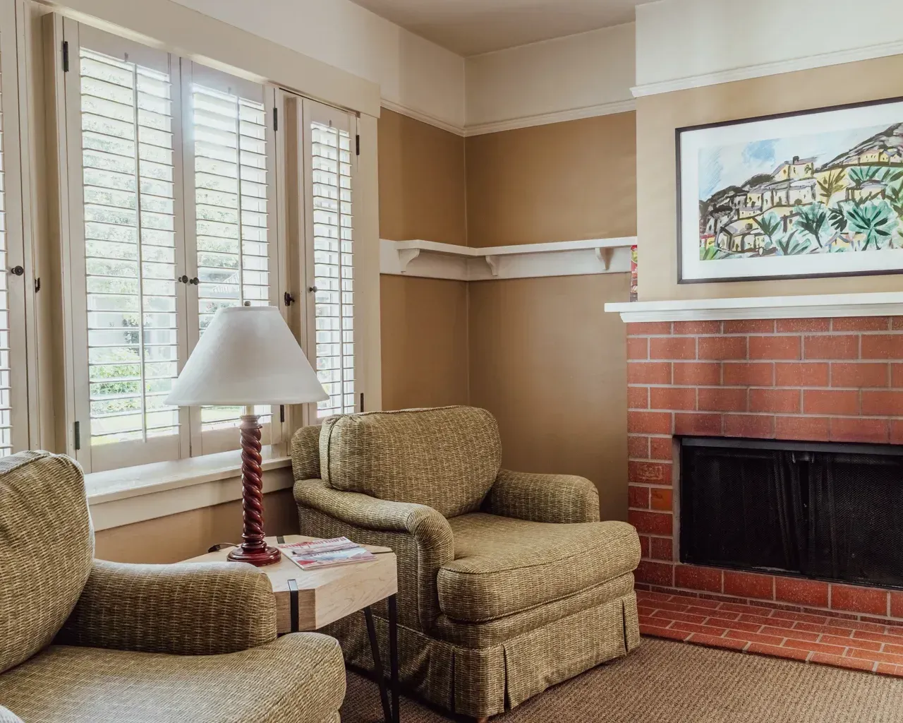 Two patterned armchairs sit by a window next to a red brick fireplace and a framed painting in a cozy living room.