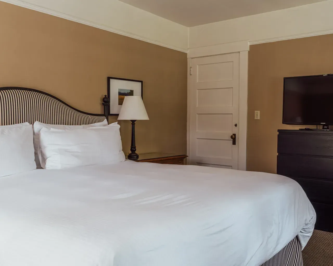 A bedroom with a bed featuring a striped headboard, white linens, a lamp on a side table, a white door, and a television.