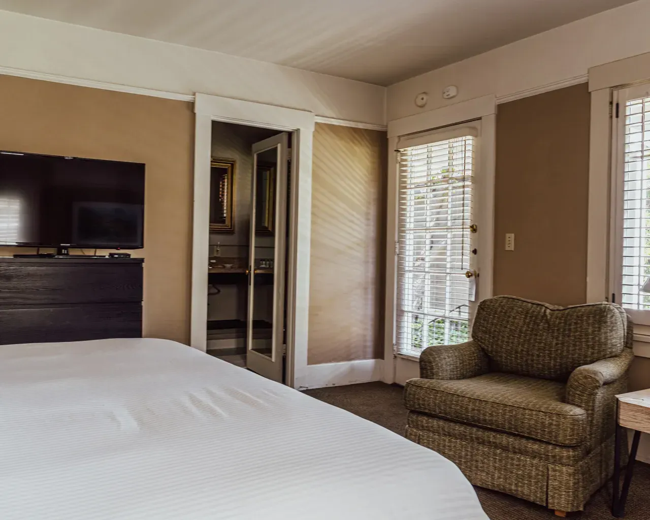 A hotel room with a bed, a TV on a dark wooden stand, an armchair, and an open doorway leading to a bathroom.