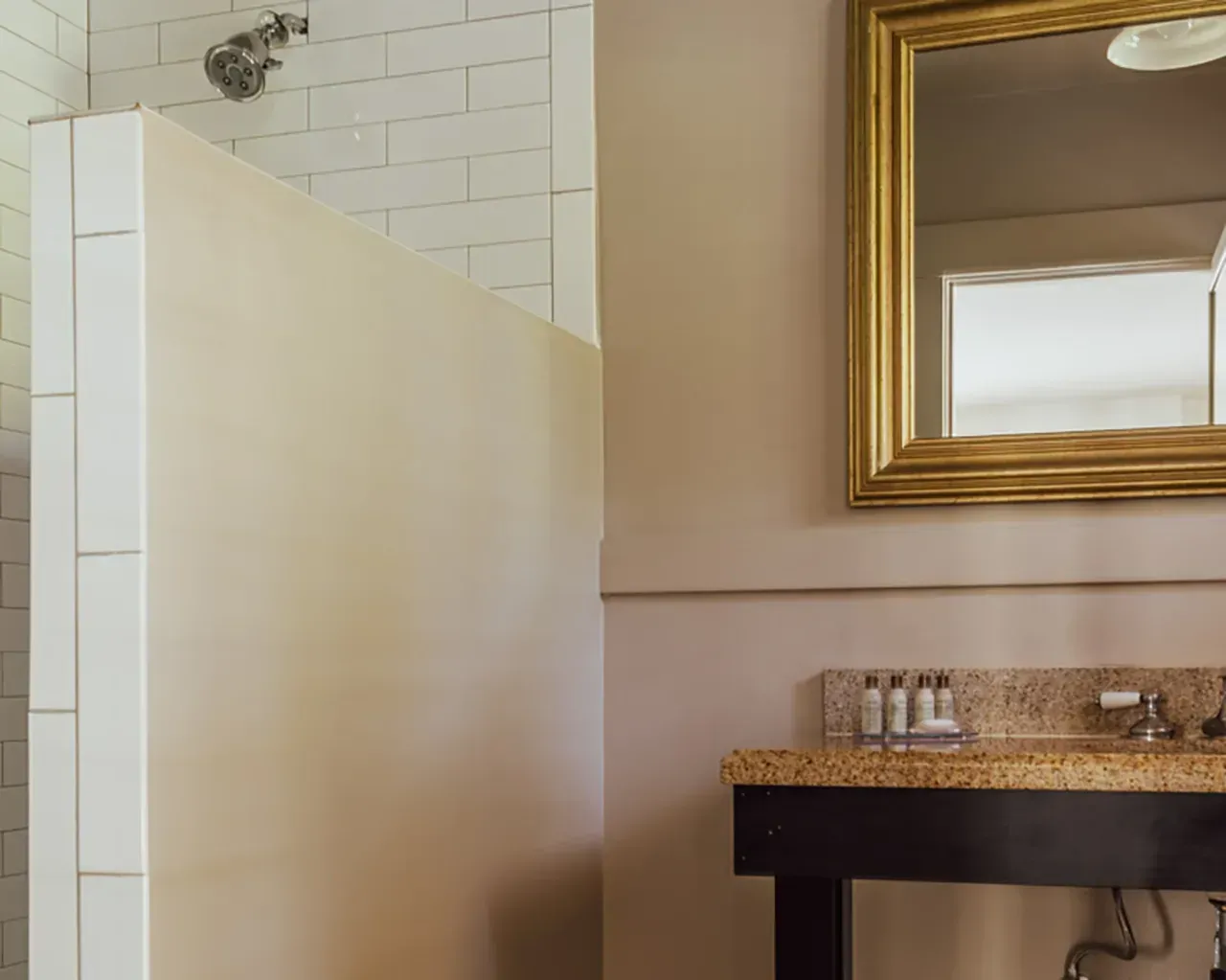 A modern bathroom features a tiled shower wall, a gold-framed mirror, and a dark wooden vanity with toiletries.