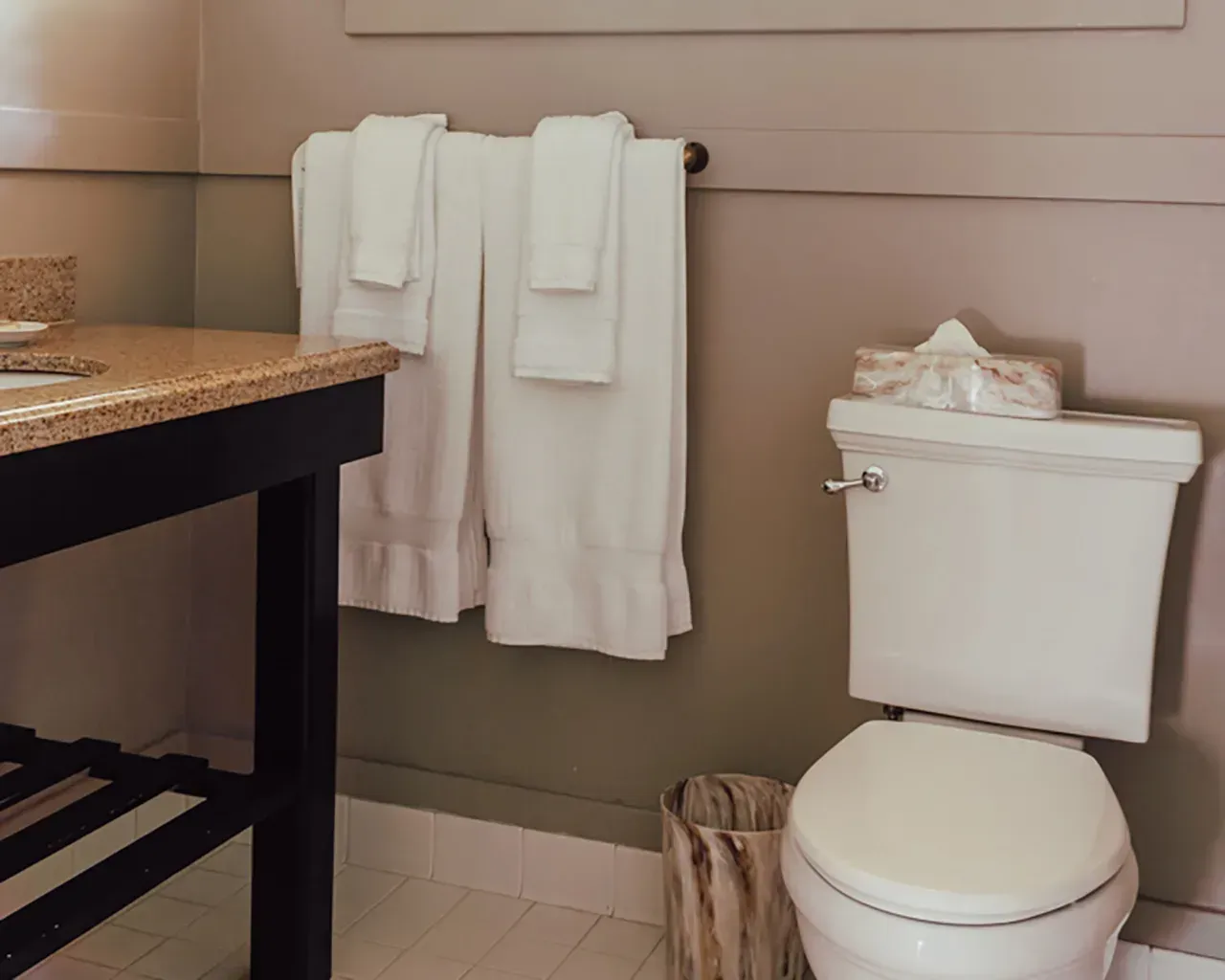 A beige-walled bathroom with a dark-legged vanity, white toilet, and two white towels hanging on a wall rack.