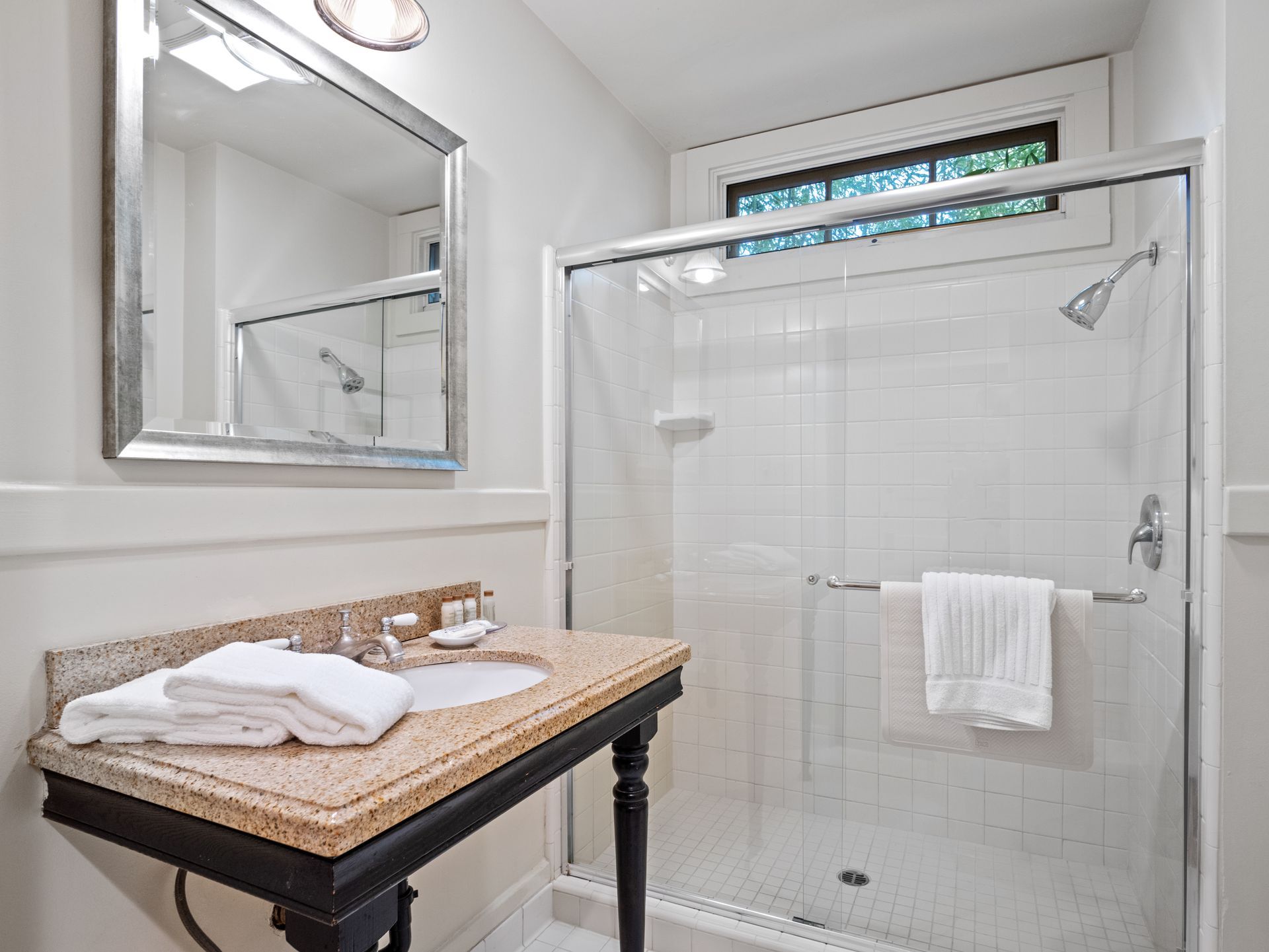 A hotel bathroom with a granite vanity, a mirror, and a walk-in glass shower with white tiled walls and a towel.