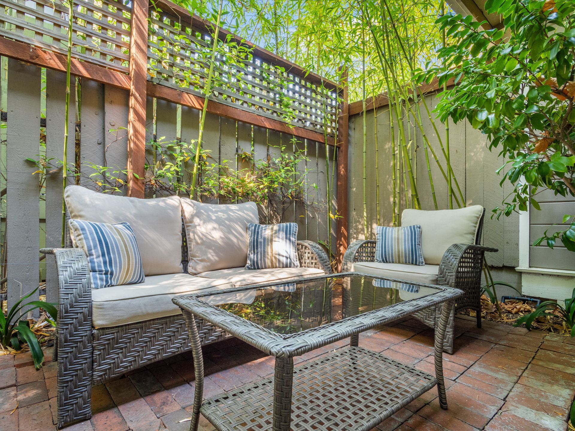 Outdoor patio with a wicker sofa, matching chairs, and a glass-topped table on a brick floor, surrounded by bamboo.