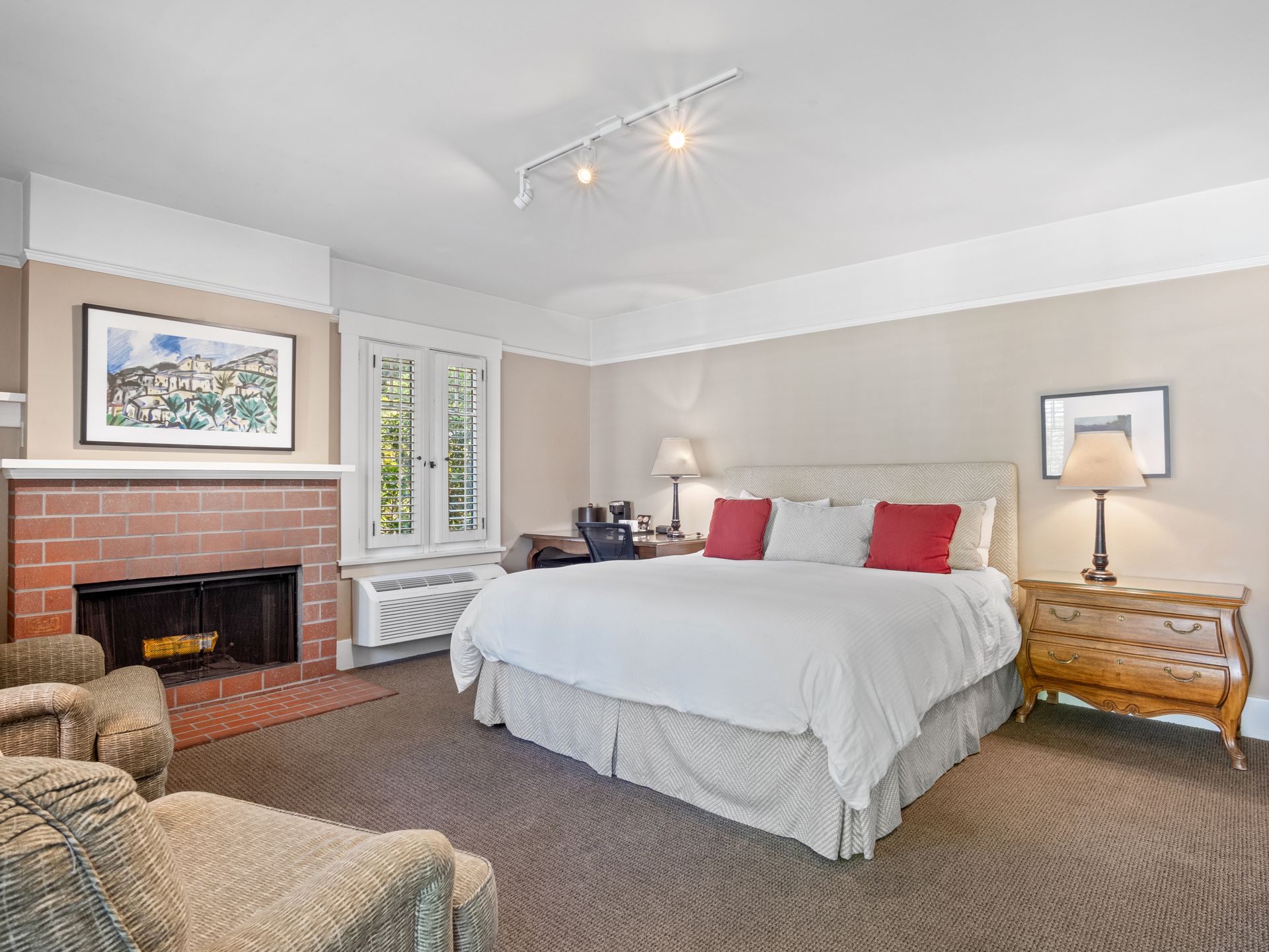 A bedroom with a king-sized bed, brick fireplace, two armchairs, a wooden nightstand, and track lighting.