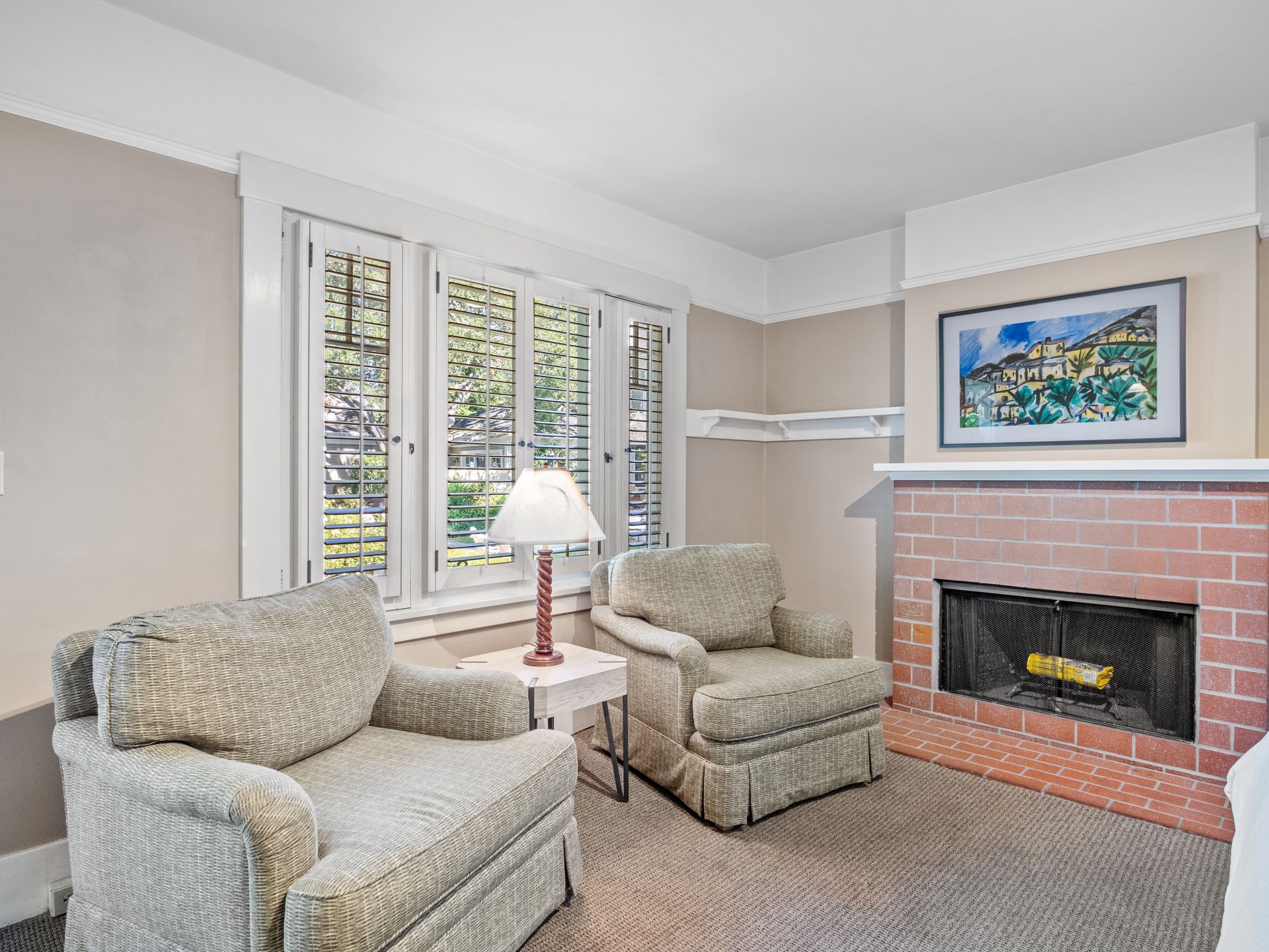 Two patterned armchairs face a brick fireplace with a framed print above it, set in a room with white-shuttered windows.