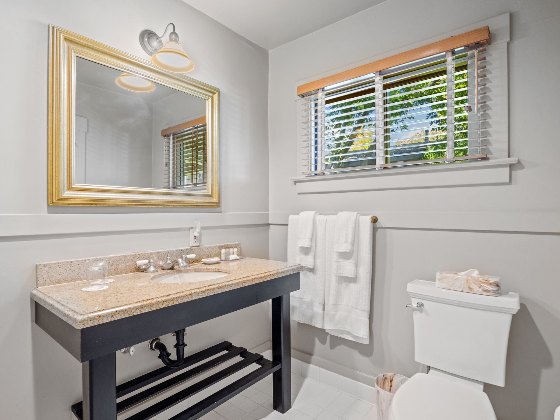 A bathroom with a vanity, sink, toilet, mirror with gold frame, and window with blinds on a light gray wall.