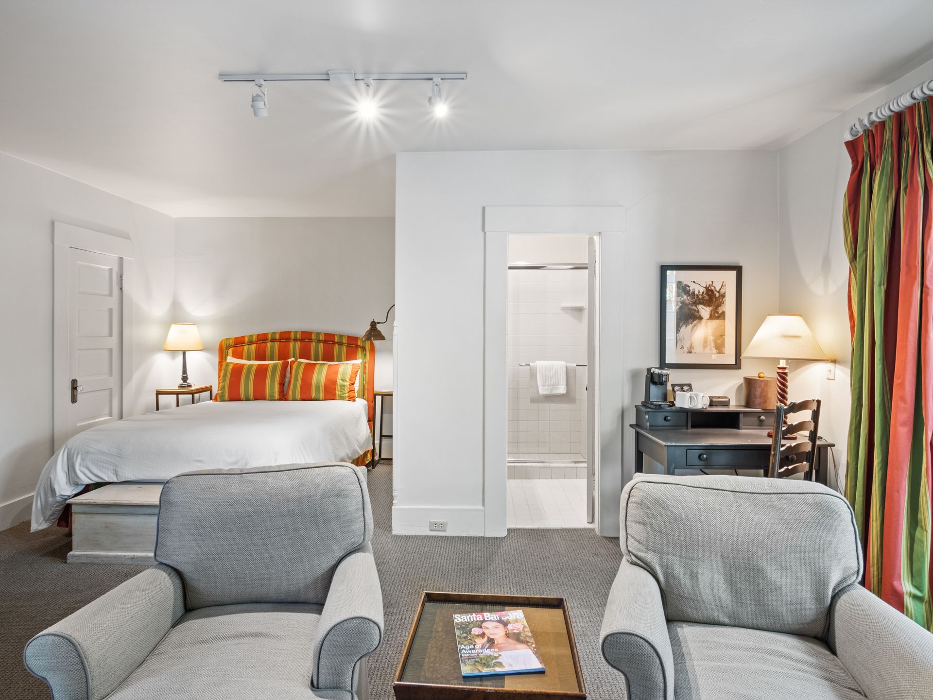 A hotel room featuring two gray armchairs in the foreground, a bed in the background, a desk, and a bathroom doorway.