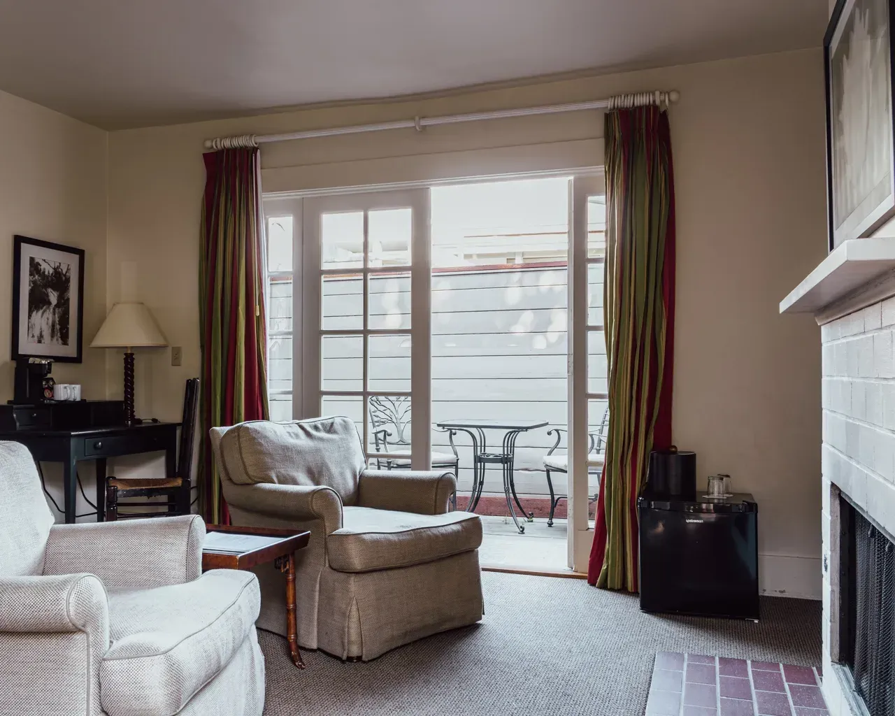 A hotel room with two armchairs, a small desk, a fireplace, and a glass door leading to a patio with outdoor furniture.