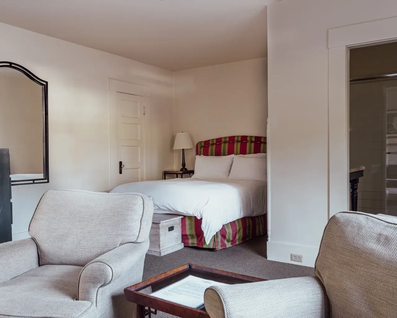 A cozy hotel bedroom with a queen bed, two light-patterned armchairs, a small table, and a mirror on a white wall.