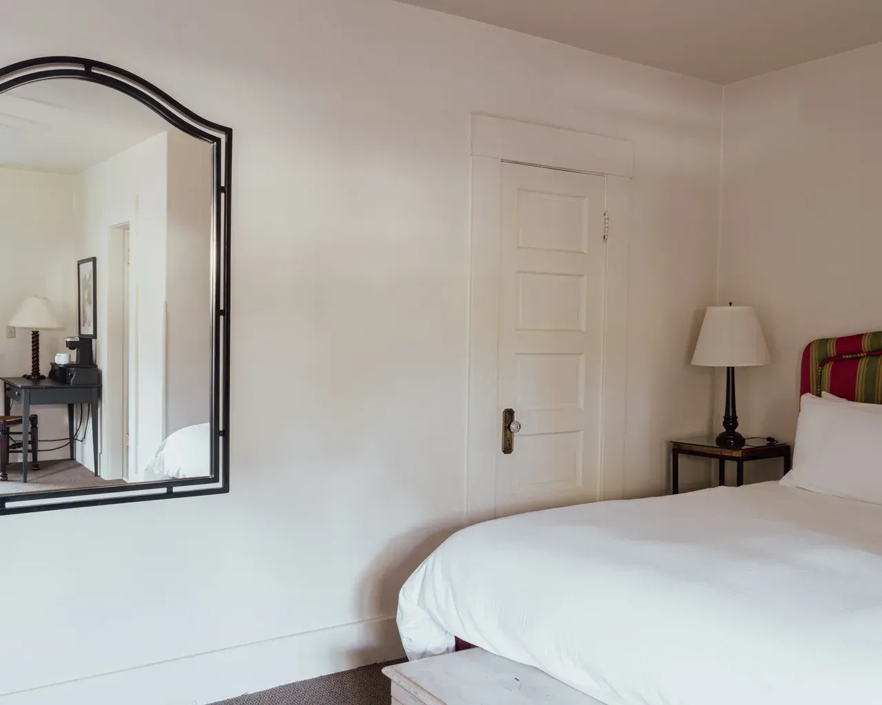 A bedroom featuring a bed with white bedding, a nightstand with a lamp, a white paneled door, and a wall mirror.