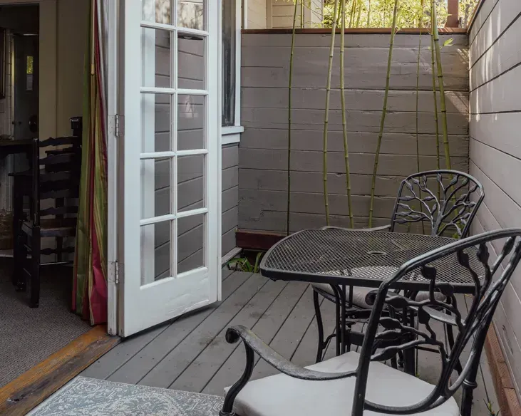 A small, gray wooden deck patio with a black metal bistro table and two chairs featuring white cushions, next to a door.