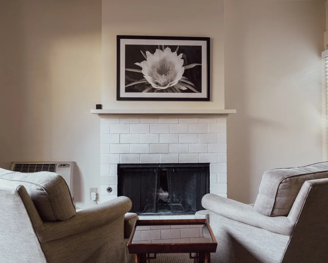 Two chairs face a white brick fireplace with a framed black-and-white flower print hanging above the mantel.