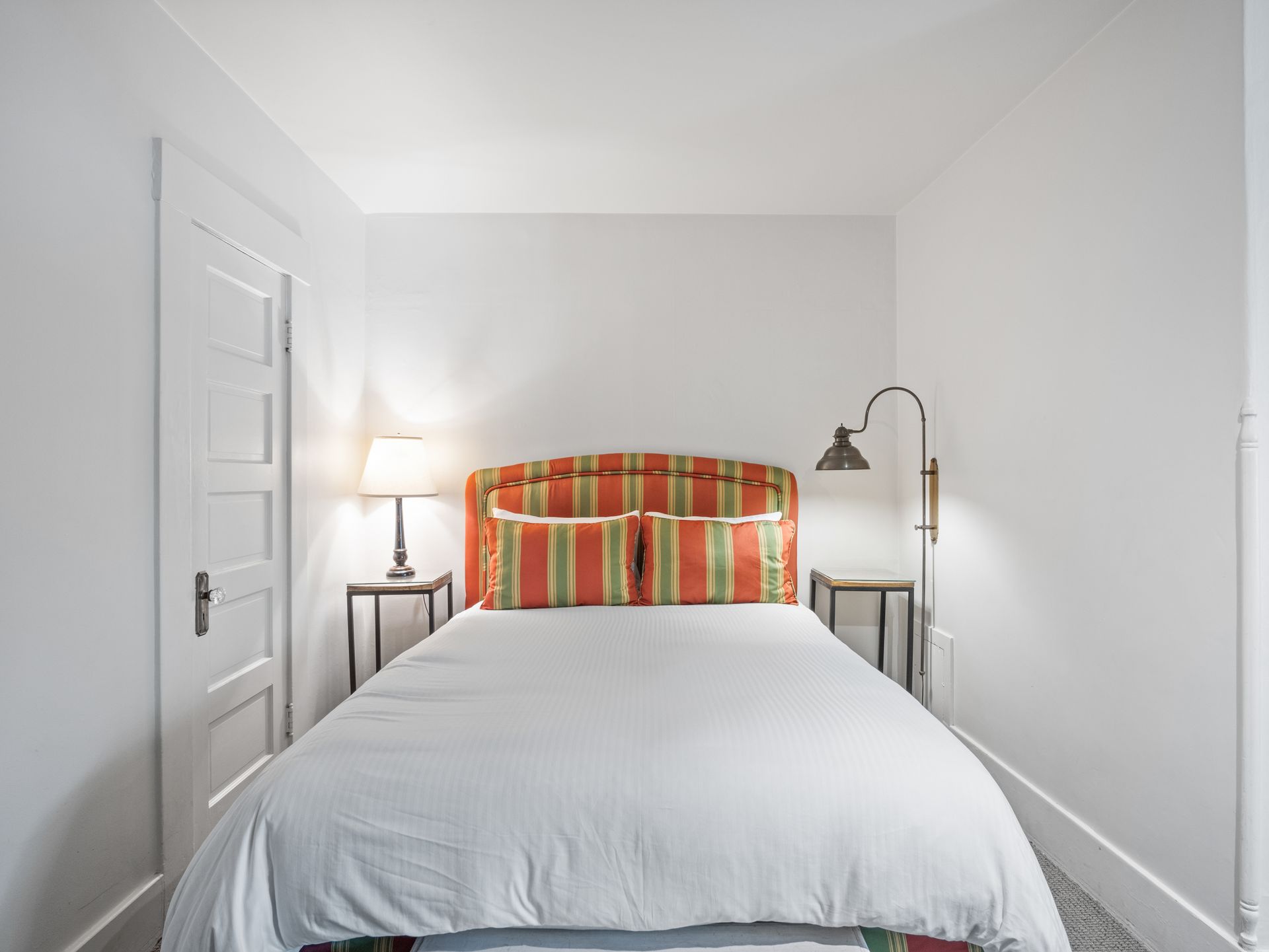 A bed with an orange and green striped headboard between two nightstands and lamps in a small bedroom with white walls.