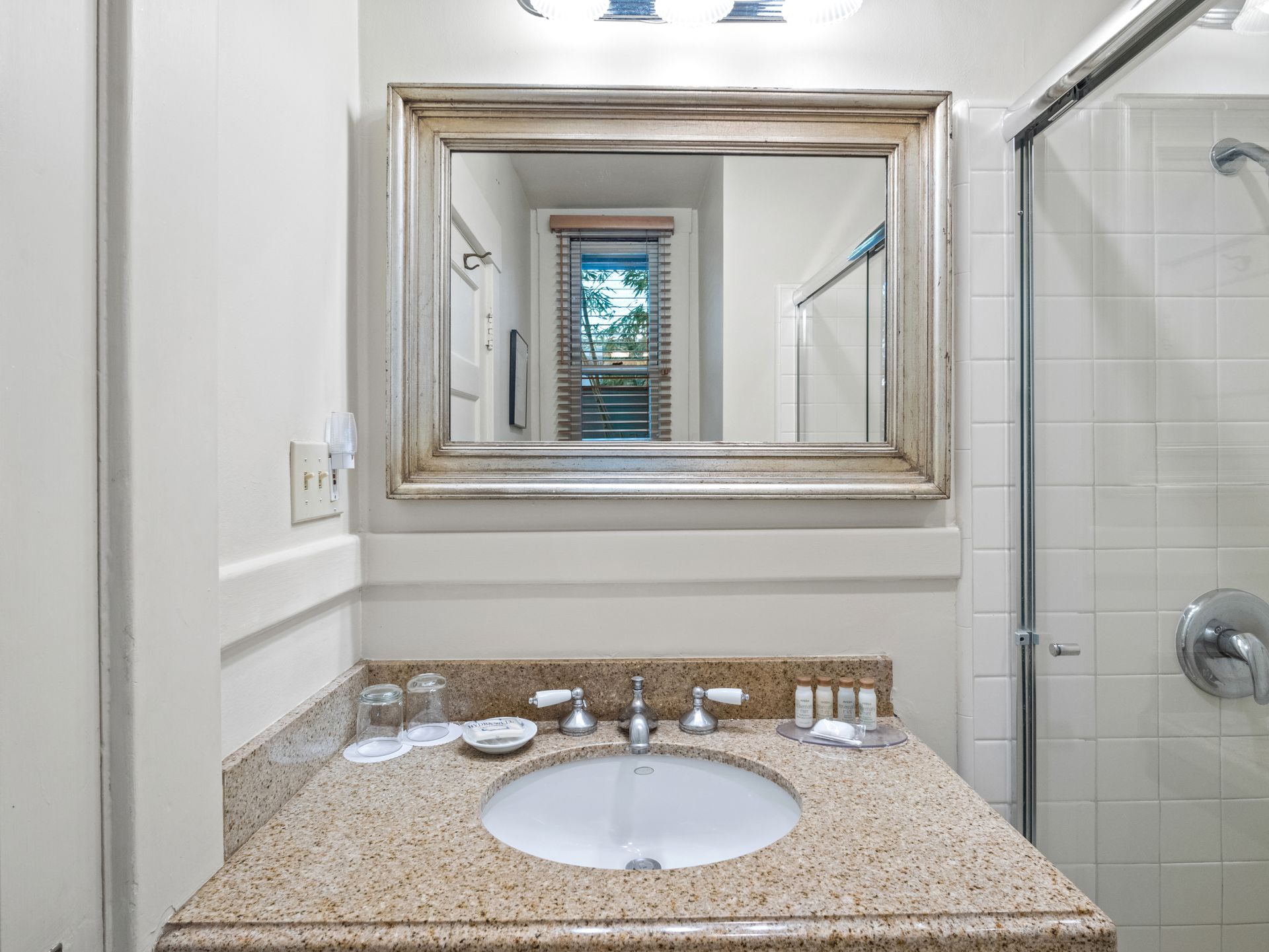 A bathroom vanity with a speckled granite countertop, white sink, decorative mirror, and an adjacent shower.