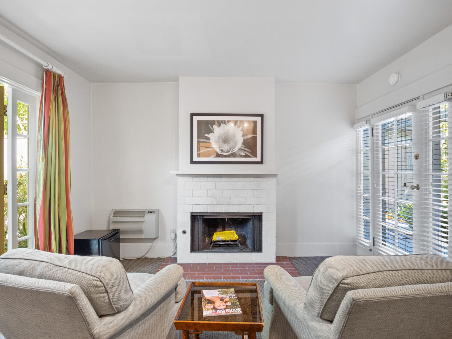 A living room features two light-colored armchairs, a small table, a white brick fireplace, and glass patio doors.