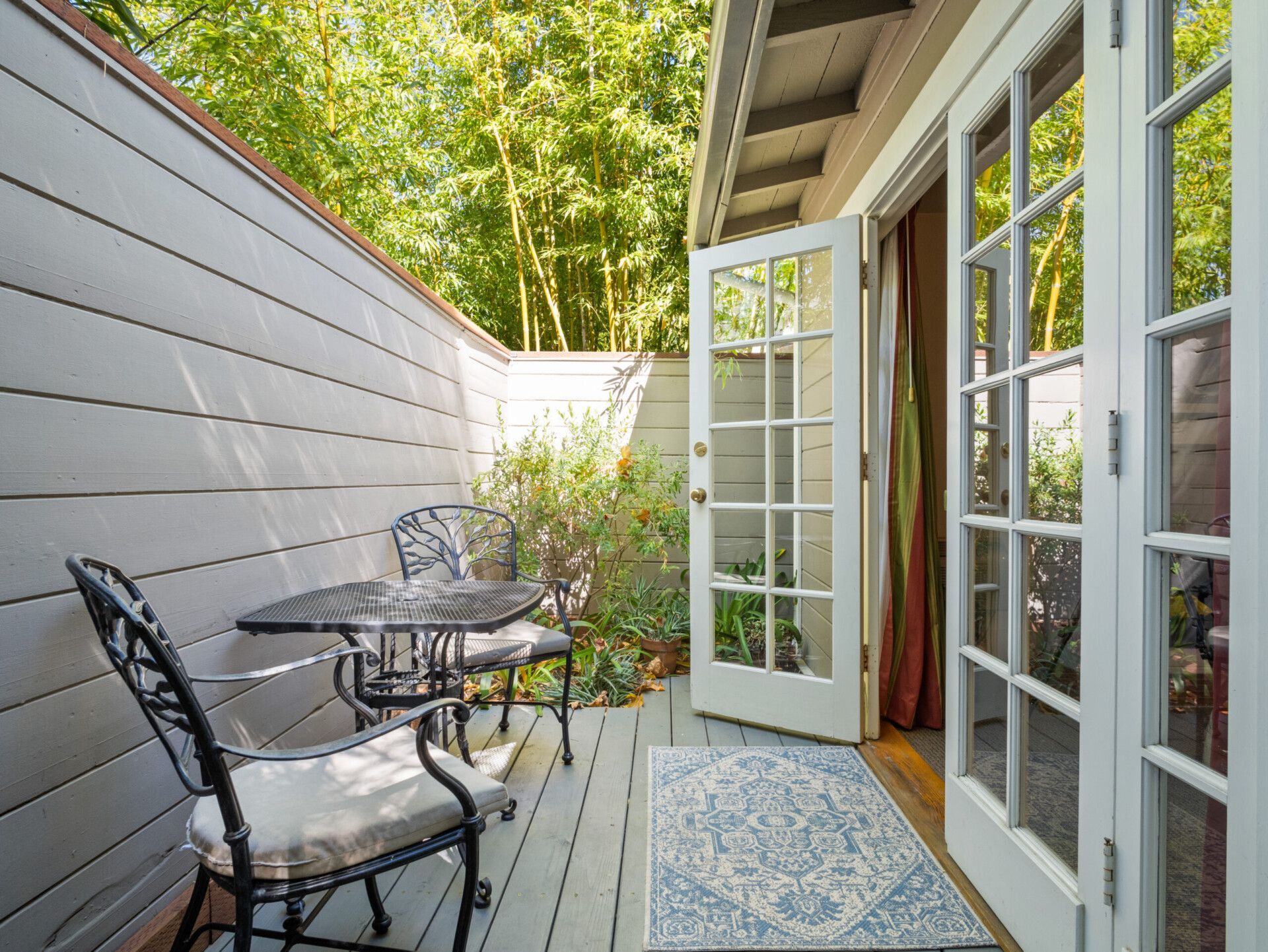 A small wooden deck patio with a metal bistro table and two chairs, next to an open white glass door leading indoors.