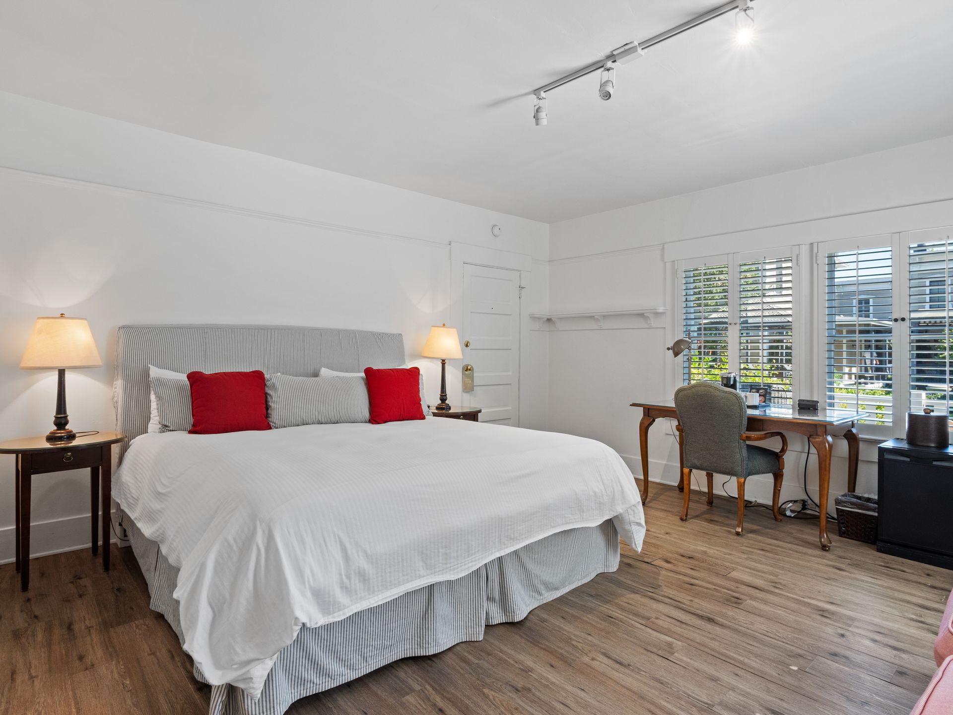 A bedroom with a king-sized bed, white bedding, red accent pillows, two side tables, a wooden desk, and a chair.