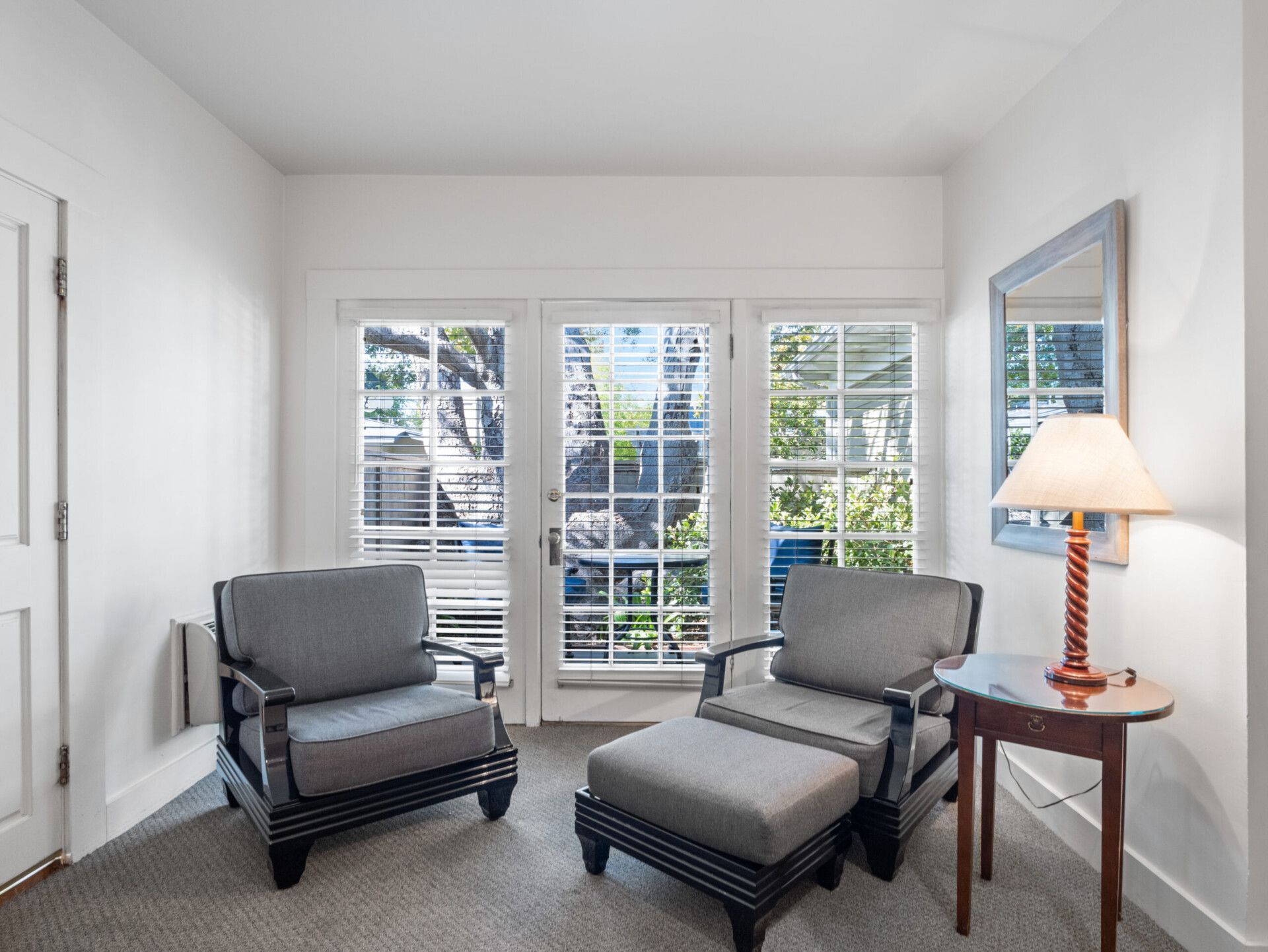 Two gray chairs, one with an ottoman, sit before glass doors in a white room with a small side table and lamp.