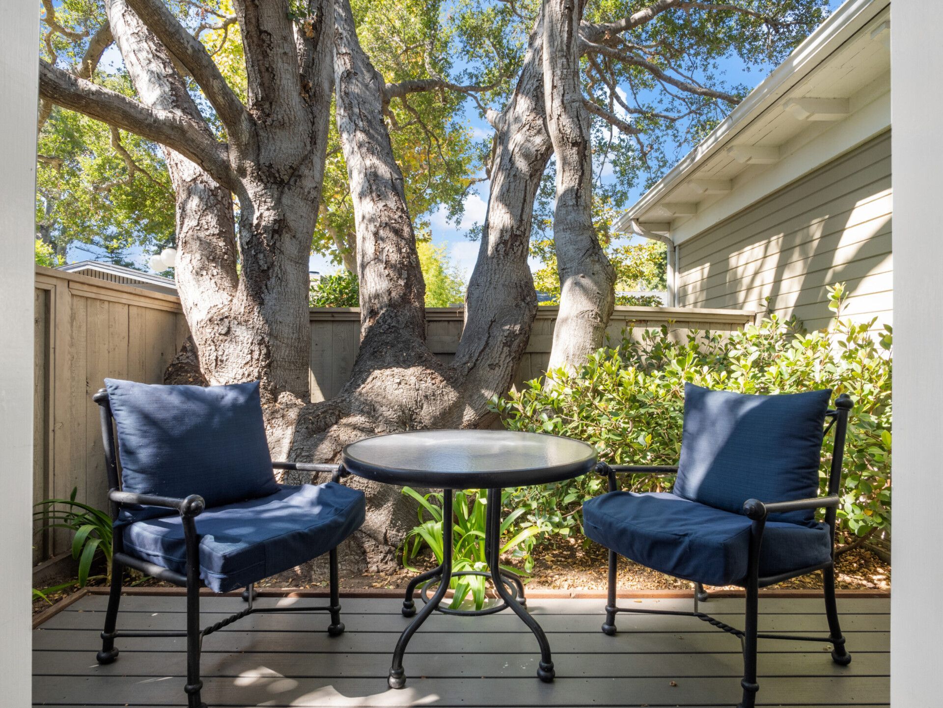 Two navy blue chairs and a small round table on a wooden patio deck against a large tree and a house exterior.