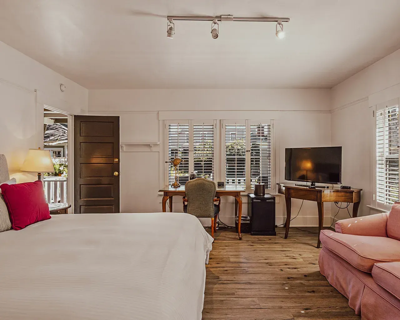 A bright, spacious room with a bed, desk, television, and small sofa, featuring hardwood floors and white walls.