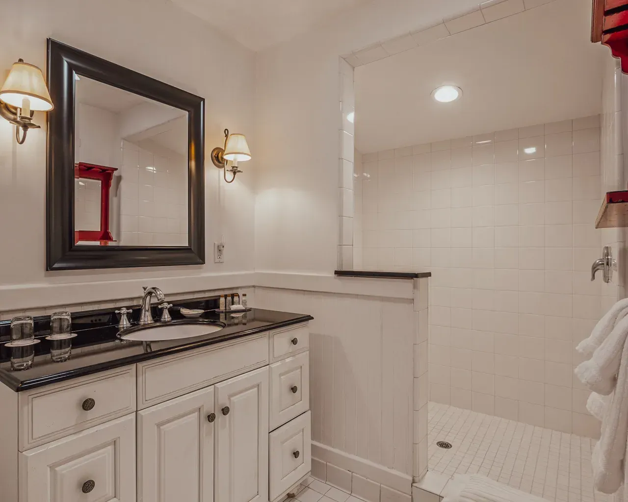 A bathroom featuring a white vanity with a black countertop, a framed mirror, wall sconces, and a walk-in tiled shower.