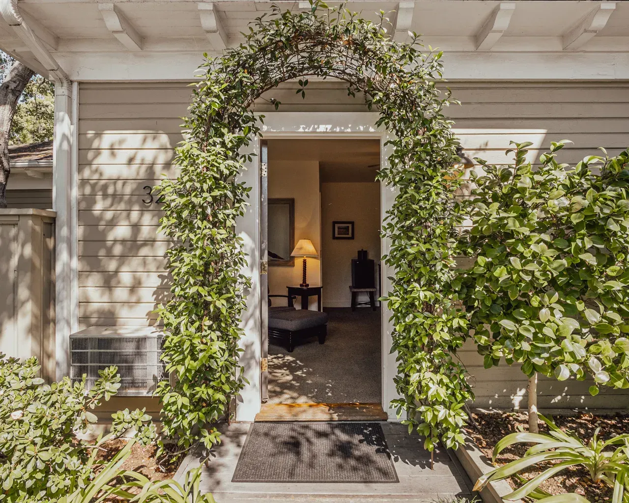 A tan wooden house entrance framed by a lush green climbing vine arch, with a glimpse into a dimly lit interior room.