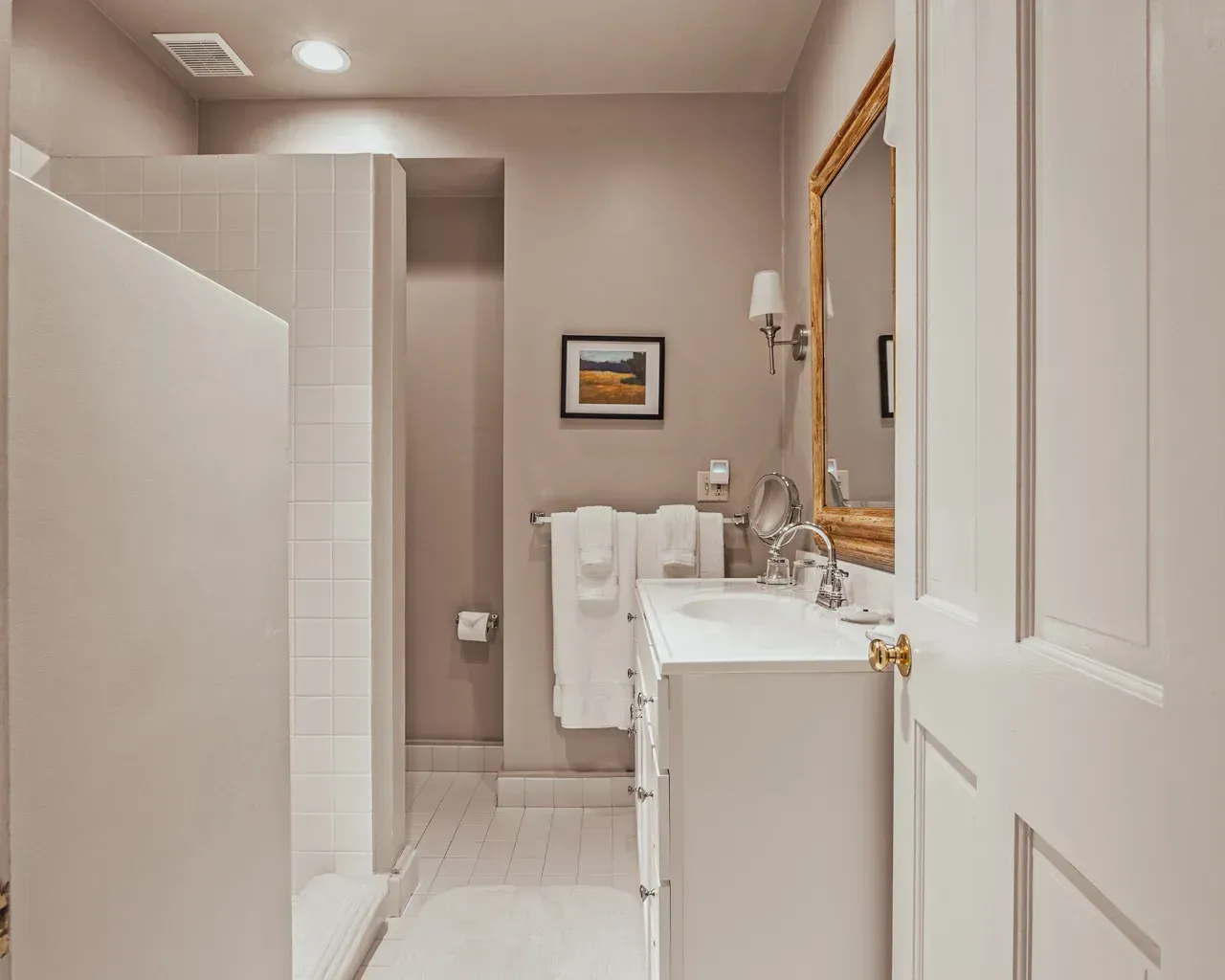 A clean, neutral-toned bathroom featuring a walk-in shower, a white vanity with a sink, and a gold-framed mirror.
