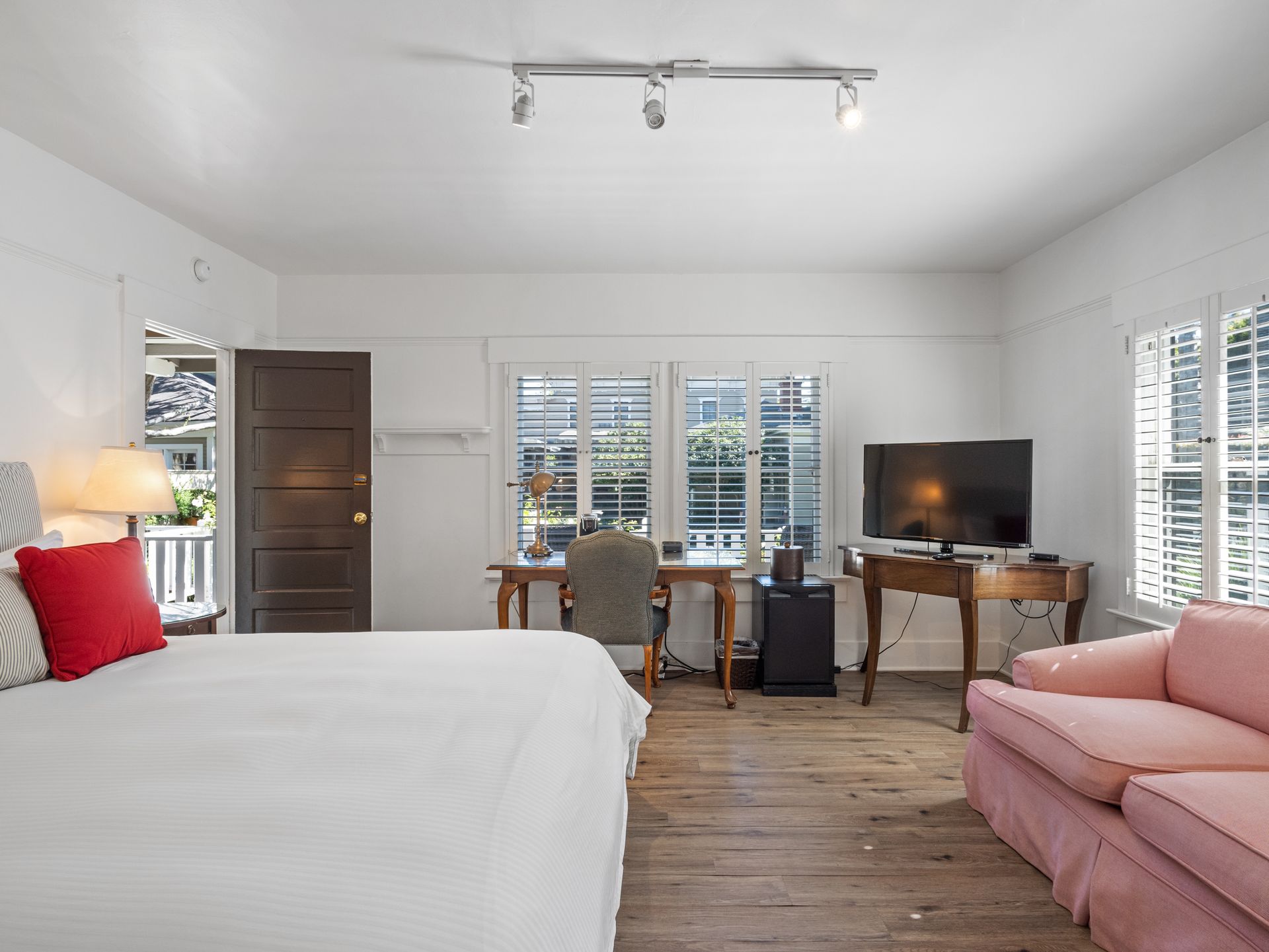 A bedroom with a large bed, pink sofa, two desks, and windows with white shutters on a wood-look floor.