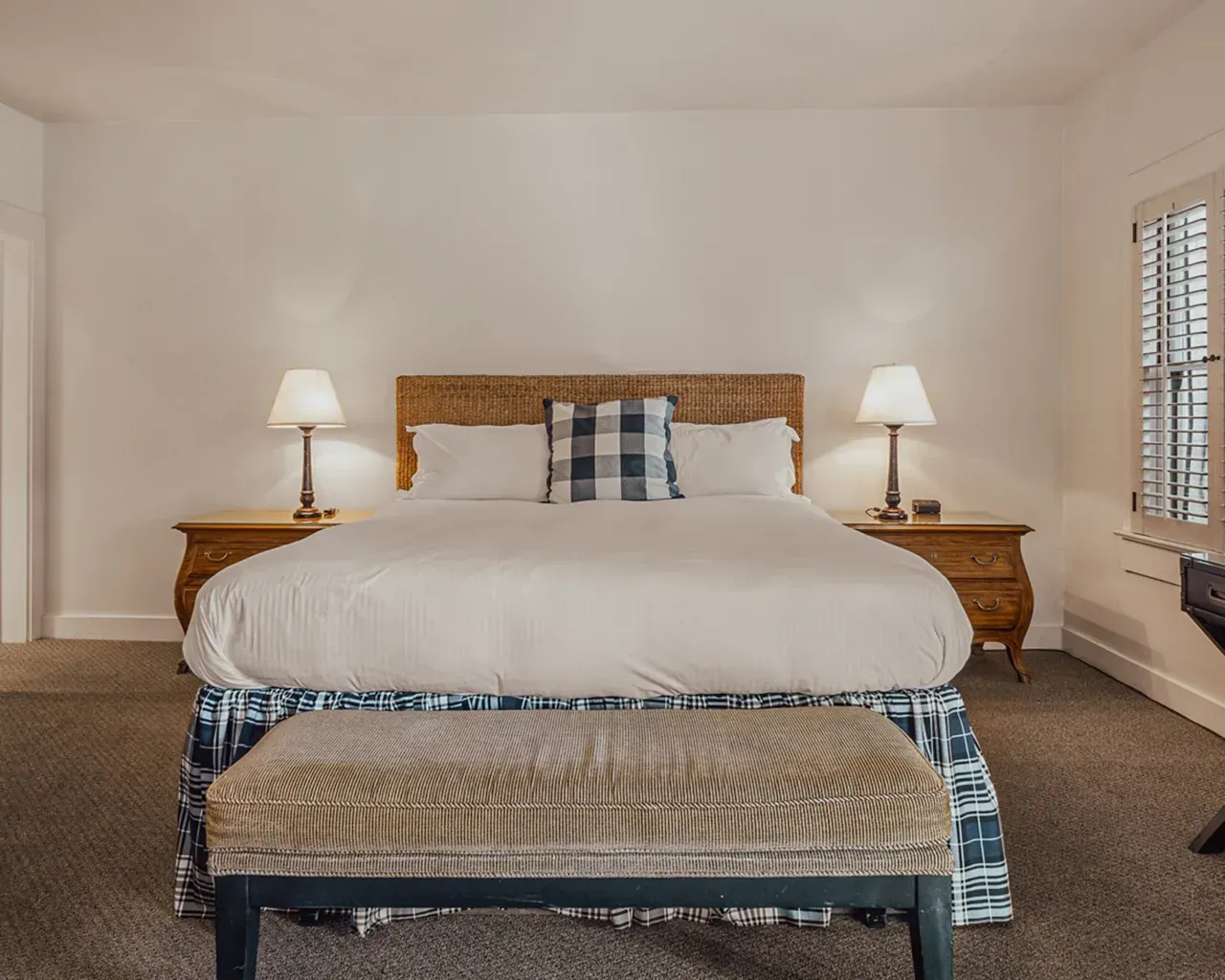 A neatly made bed with white linens and a plaid accent pillow sits between two wooden nightstands with lamps.