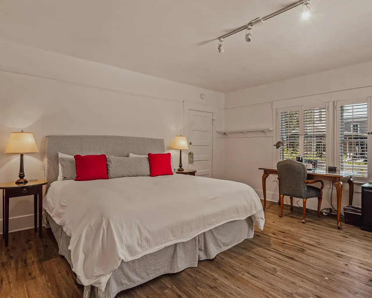 A hotel bedroom with a large bed, striped headboard, white linens, two red pillows, nightstands, and a desk by a window.