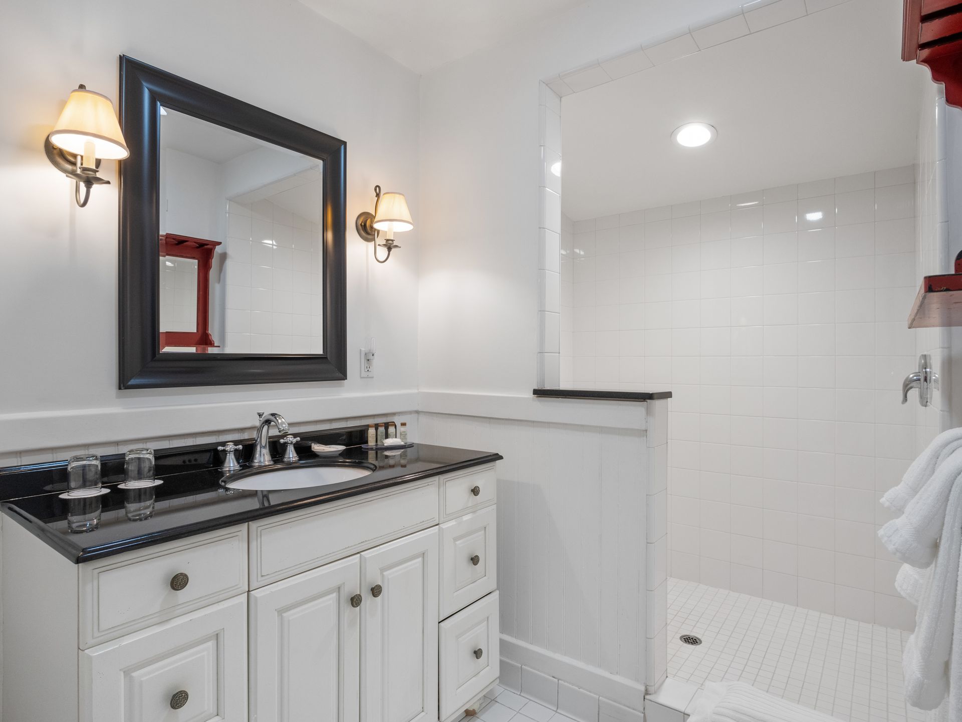 A white bathroom vanity with a black countertop and mirror, next to a white-tiled walk-in shower.