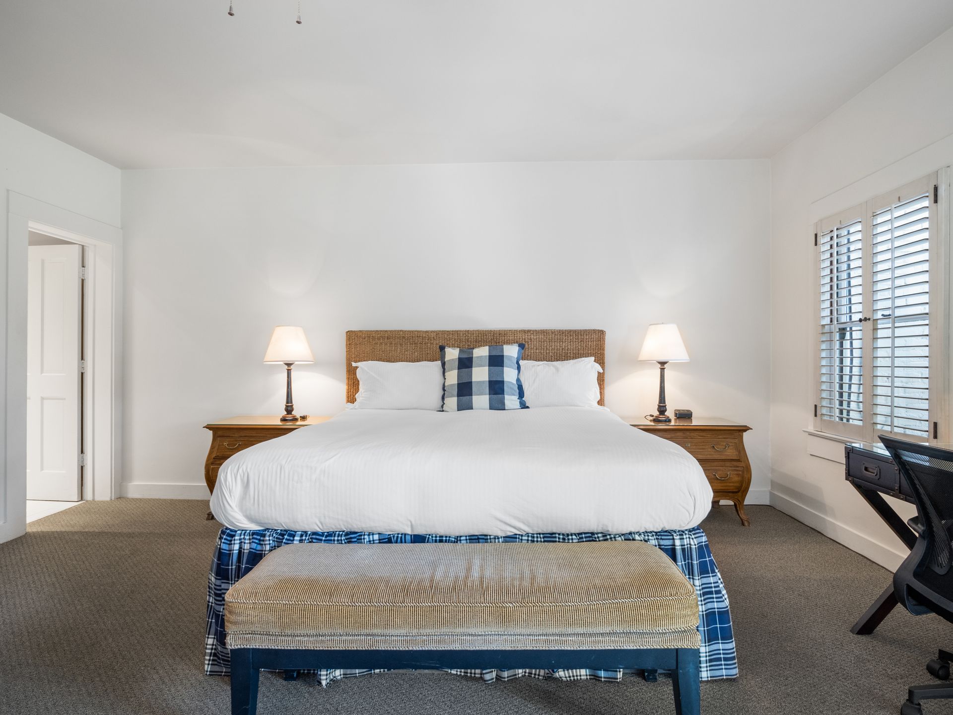 A bright bedroom with a king-sized bed, woven headboard, white bedding, two bedside lamps, and a patterned bench.