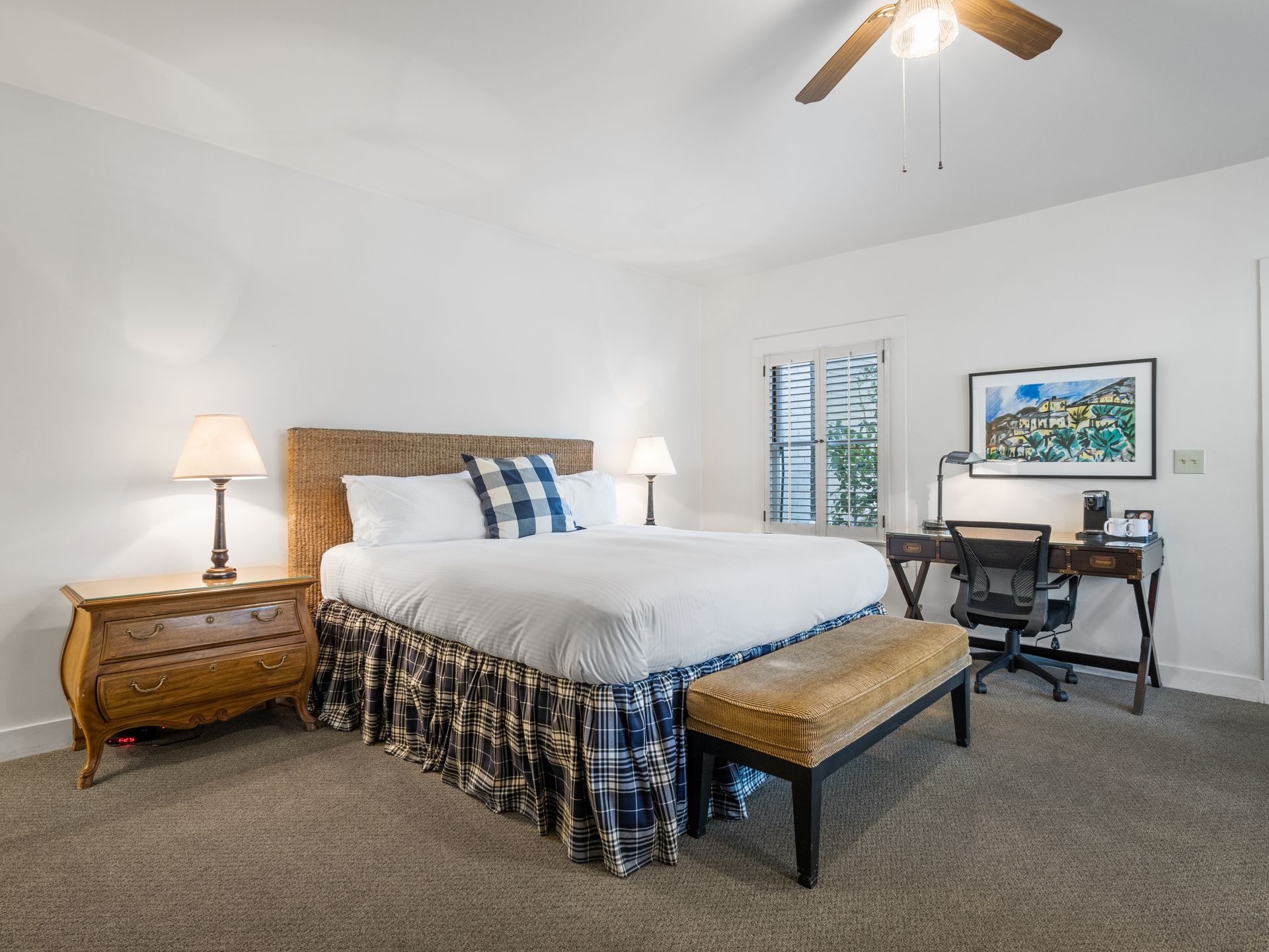 A bedroom with a wicker-framed bed, blue plaid pillows, a wooden nightstand, and a desk with a chair under a ceiling fan.