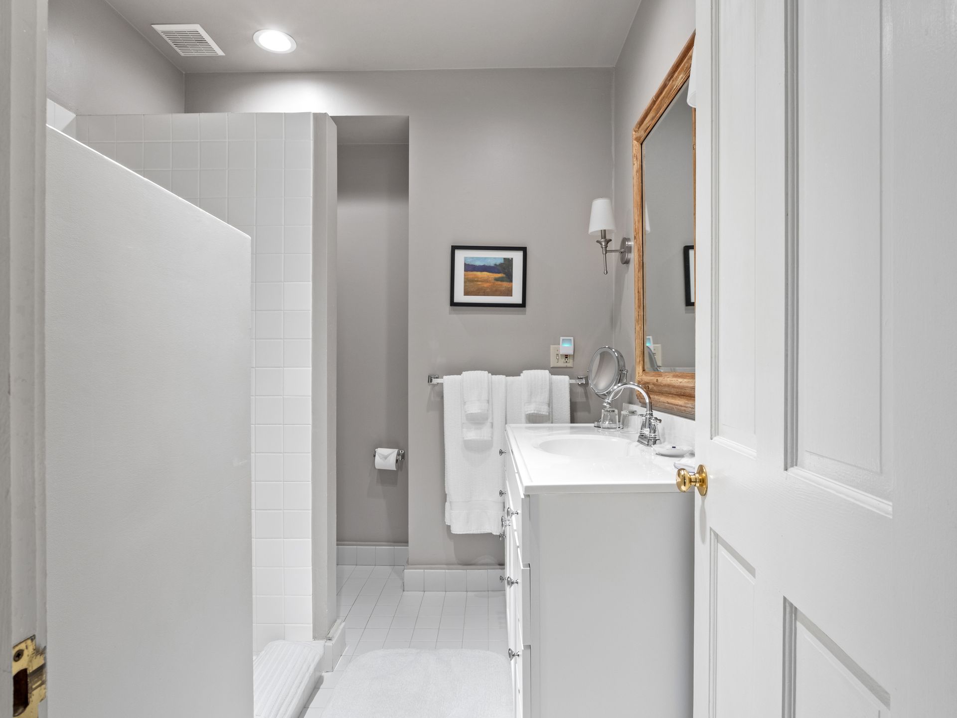A bright, modern bathroom with a white vanity, a tiled walk-in shower, grey walls, and a wood-framed mirror.