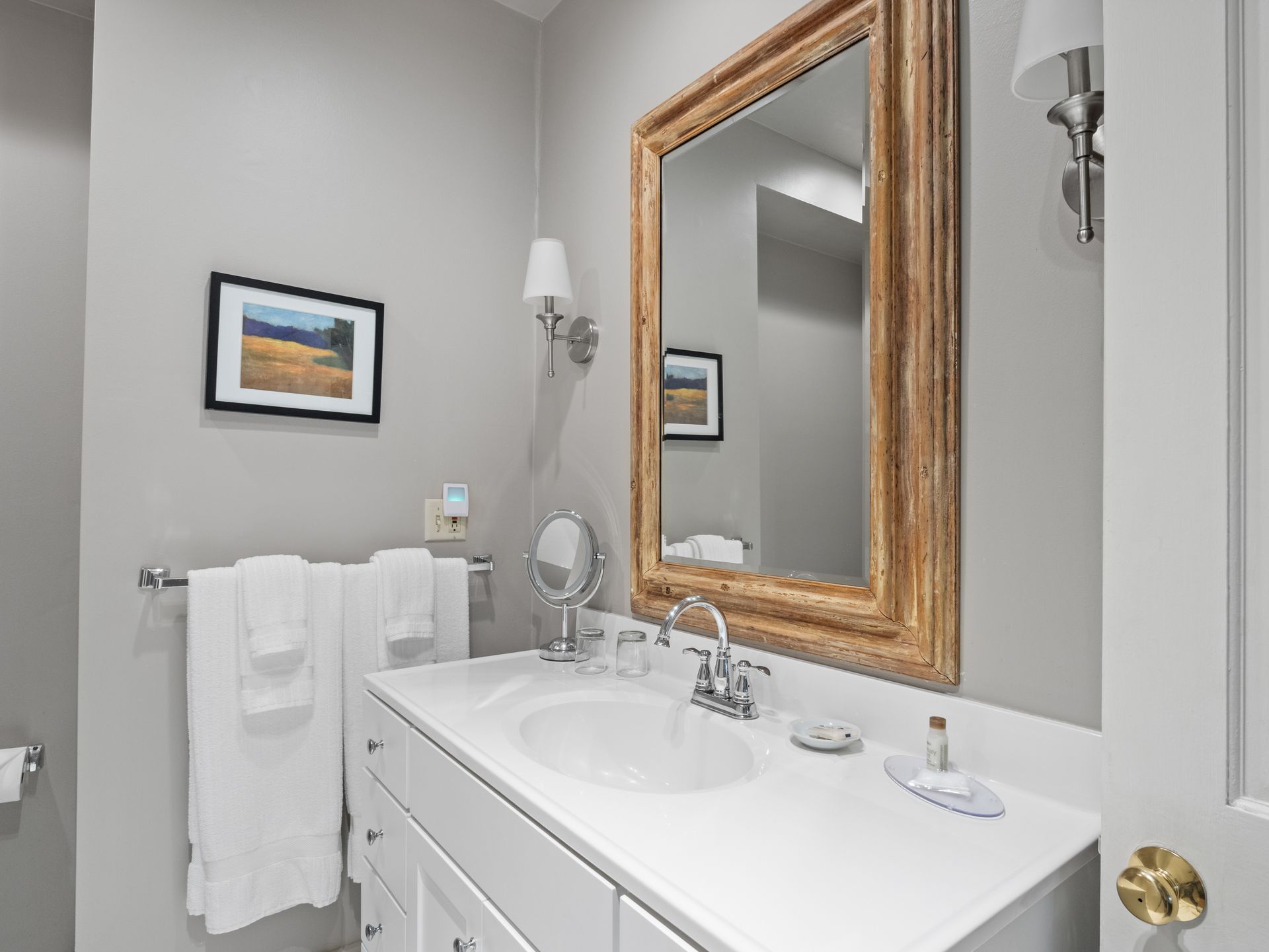 A modern, neutral-toned bathroom featuring a white vanity, a gold-framed mirror, and a towel rack with white towels.