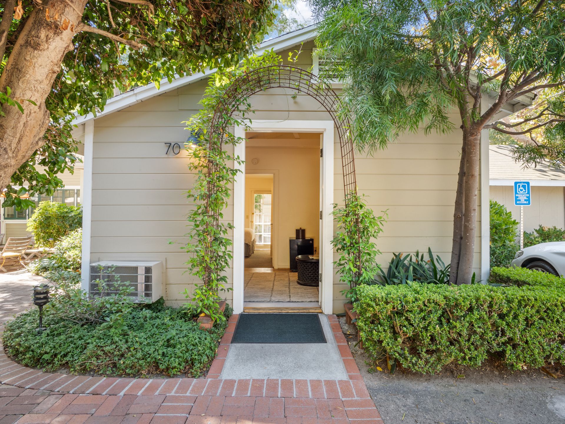 A light-colored building entrance featuring a decorative metal arch covered in vines, surrounded by trees and shrubs.